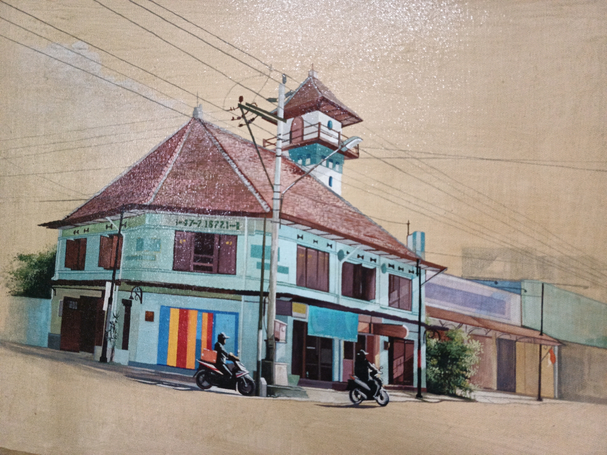 This is a painting depicting a street scene in Pabelan, Indonesia. The focal point is a two-story building with a prominent, multi-tiered roof, painted in light blue and adorned with dark red shutters. The building's facade displays a series of windows and doors, with a vertical arrangement of colorful stripes (red, yellow, blue) on the lower left side. A utility pole with wires is positioned in front of the building. Two individuals on motorcycles are present: one on the left, riding a black motorcycle with an orange box on the back, and another on the right, riding a white motorcycle. The street in front of the building is paved and slopes slightly upwards. To the right of the main building, a lower structure with awnings extends along the street, featuring more doors and windows. Green foliage is visible behind the awning of this extension. The background consists of a neutral-toned sky with visible overhead wires. The painting style is detailed, with a textured canvas evident.