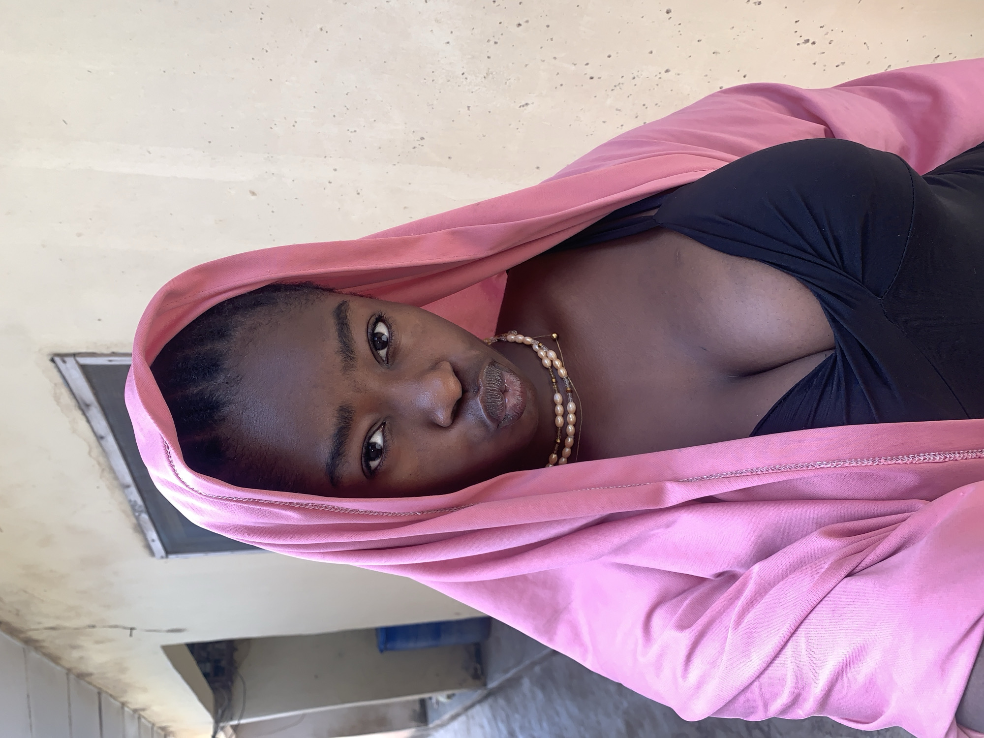 A woman with dark skin is shown wearing a black top and a pink hooded garment. She has a pearl necklace and is looking directly at the camera. The background suggests an interior space, with a beige ceiling and a glimpse of what might be a window or vent. The location is reported to be Rafin Par, Nigeria.