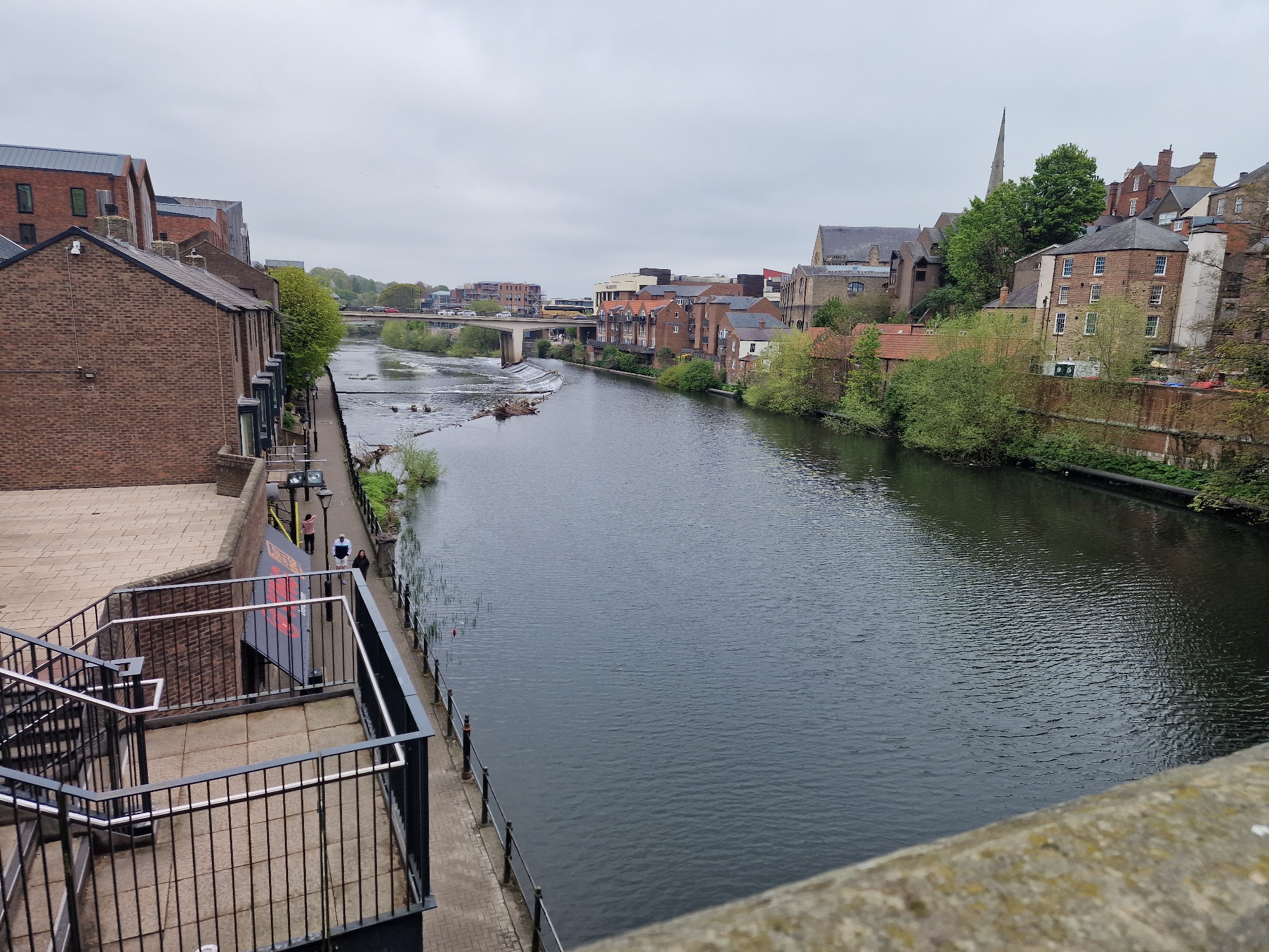 The image shows a waterway view of the River Wear in Durham, United Kingdom, under an overcast sky. The river flows from the middle ground towards the foreground. Buildings and trees line the banks on both sides. A concrete bridge spans the river in the middle ground with traffic on it. A weir is visible in the river. On the left side of the image is a brick building with a staircase and railings leading down towards the river level, a sign is hanging on the brick wall. Three people are walking on the pathway adjacent to the railing. The foreground shows the top of a stone bridge from which the image was taken.