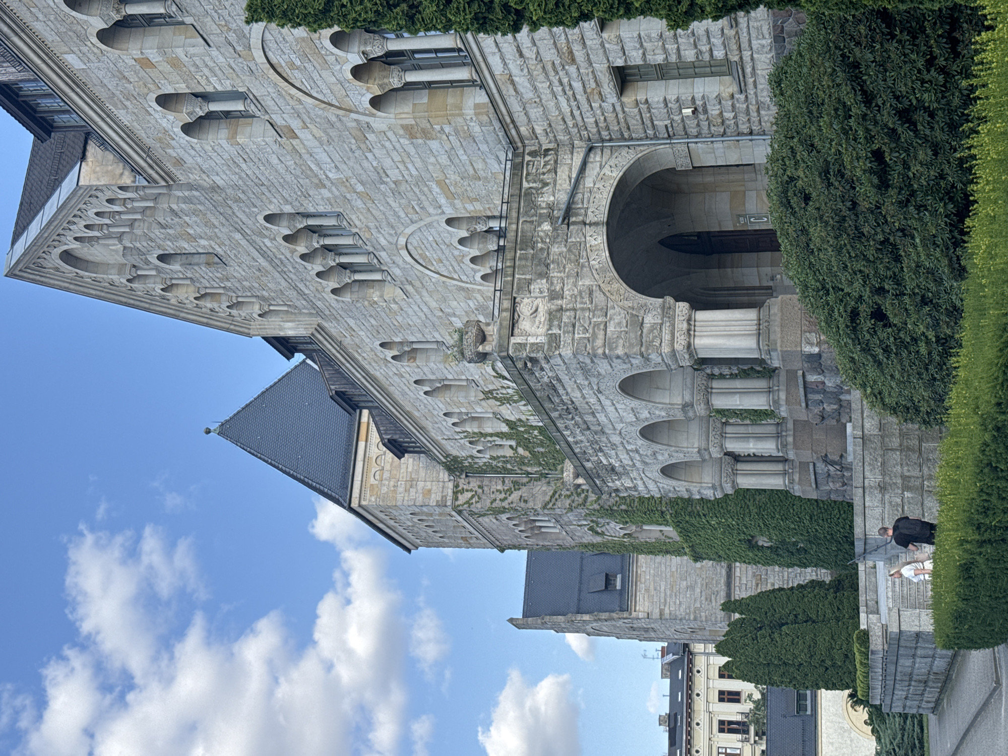 The image depicts the historic Imperial Castle in Poznan, Poland. The castle, constructed from light-colored stone, features a large tower with a dark, pointed roof. Green ivy covers portions of the castle walls, particularly around the tower and the main facade. The castle exhibits Romanesque and Gothic architectural elements, including arched windows and decorative stonework.
In the foreground, a pathway leads up to the castle entrance, flanked by manicured green bushes and hedges. Two people, a woman and a man, are walking up the stone steps towards the castle. The woman, wearing a white top and light-colored skirt, is slightly ahead of the man, who is dressed in a black t-shirt and dark pants. They appear to be engaged in conversation.
The sky above is a clear blue with scattered white clouds. To the left of the castle, other buildings can be seen, suggesting an urban setting surrounding the historic landmark. The overall scene conveys a sense of historical grandeur and a peaceful, sunny day.