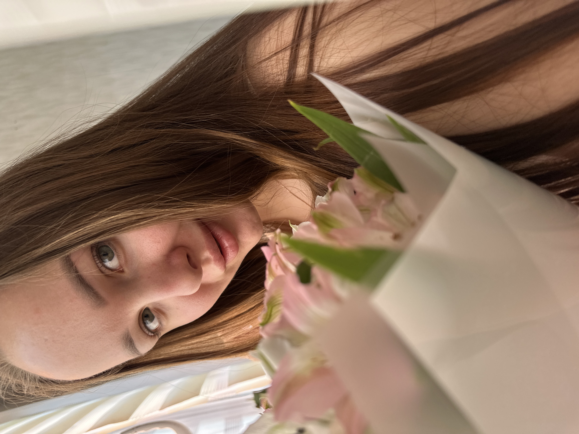 A young woman with light brown hair and fair skin poses with a bouquet of pale pink flowers wrapped in white paper. Her face is tilted towards the camera, revealing her eyes and lips. The background appears to be a light-colored wall or surface. There is no definitive evidence to confirm the location is in Chaikovsky, Russia.