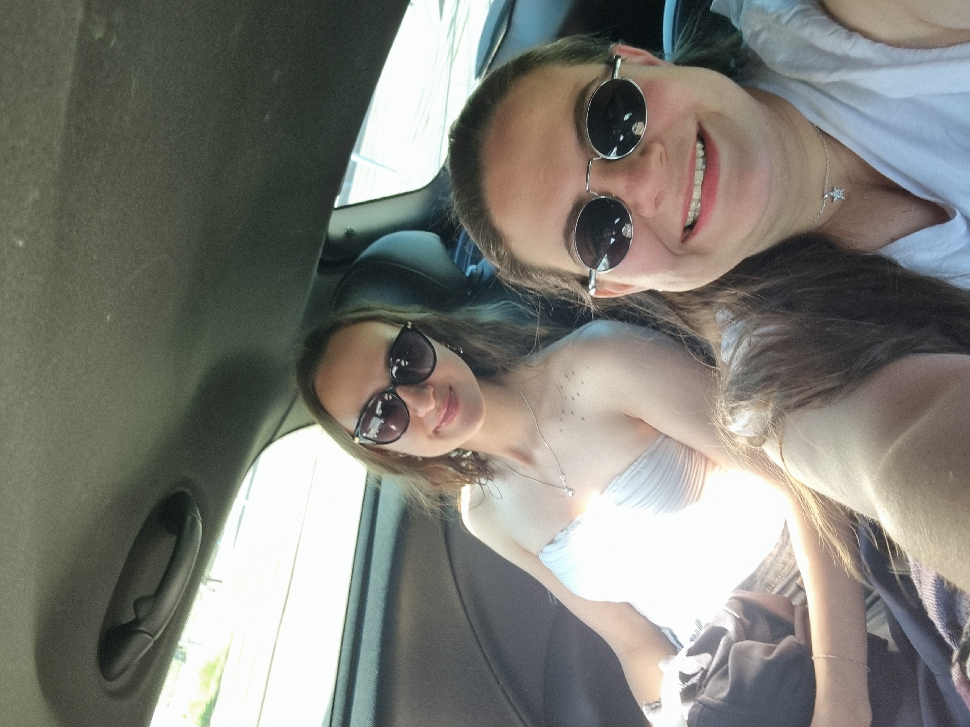 Two women are posing in a car. One woman is wearing round sunglasses and a white shirt, and is smiling. The second woman is wearing dark, oval sunglasses and a white strapless dress. The car is interior is dark and the sun is shining through the window. The photo was likely taken in a city but does not contain enough information to ascertain if the location is Saarbrücken, Germany.