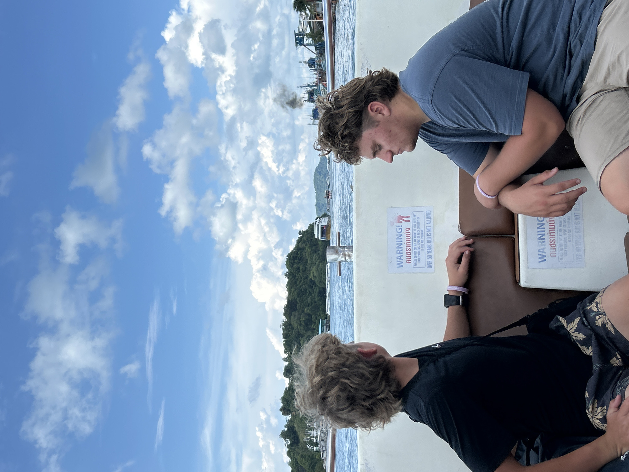 Two young males are seated on a boat with brown leather-like seating, facing away from the camera and looking out at a body of water. The male on the left has light brown, curly hair and is wearing a black t-shirt and patterned shorts. He has a black smartwatch on his left wrist and a purple wristband on his right wrist. The male on the right has dark brown hair with a mullet hairstyle and is wearing a blue t-shirt and tan shorts. He also has a purple wristband on his left wrist.
In the background, there is a green, tree-covered island on the left and a harbor with several boats and a building on the right. Black smoke is emanating from one of the boats in the harbor. The sky is blue with scattered white clouds. There are two white signs with blue text affixed to the back of the seats in front of the males. The text on the signs appears to be in English and Thai. One sign reads "WARNING" in English, followed by Thai text and then "PLEASE DO NOT SIT AT THE FRONT IF YOU HAVE ANY BACK PROBLEMS, SPINE OR NECK SURGERY OR ARE PREGNANT. OVER 50 YEARS OLD IS NOT ALLOWED." The location is identified as Ratsada, Thailand.