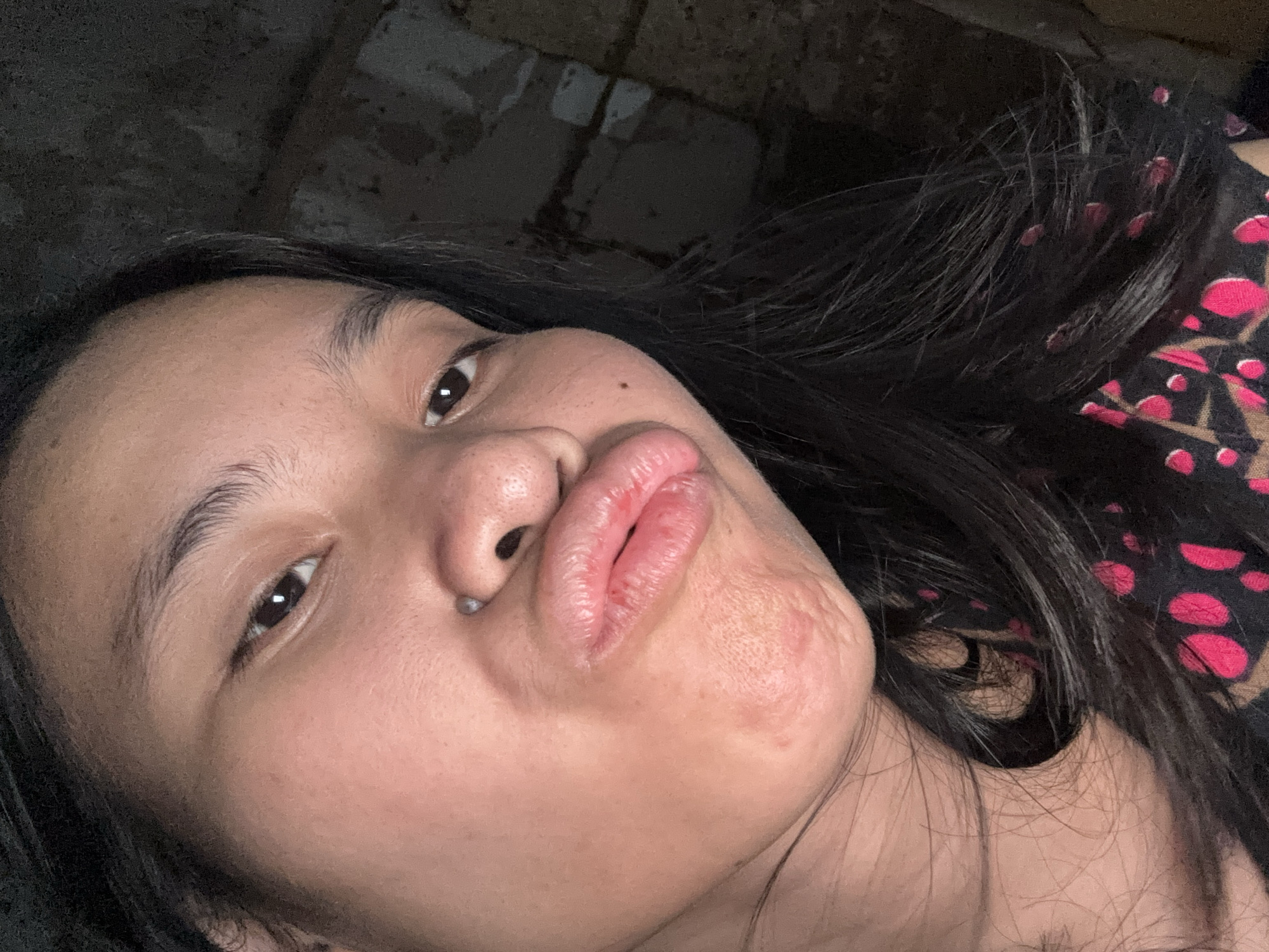 The image is a close-up selfie of a young woman with dark hair, wearing a black top with a pink and brown spotted pattern. She is making a puckered face towards the camera. A silver piercing is visible on her nose. The background is dark and appears to be an indoor environment with textured, possibly brick or concrete walls. The lighting is dim, creating shadows and highlighting her facial features. There is no identifiable location in East Jakarta, Indonesia, present in the image.