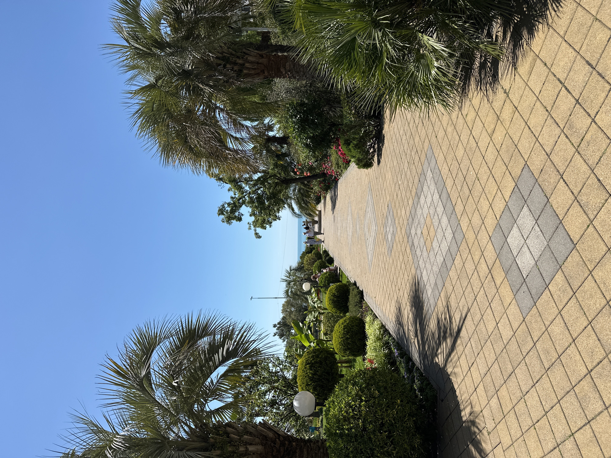 The image shows a walkway in Sochi, Russia. The walkway is paved with tan and gray diamond-patterned tiles and curves gently towards the horizon. Along the sides of the walkway, there are neatly manicured bushes and palm trees. In the distance, people are visible near the sea. The sky is clear and blue, suggesting a sunny day. White globe lights are placed among the bushes. Shadows from the palm trees fall across the tiled walkway.