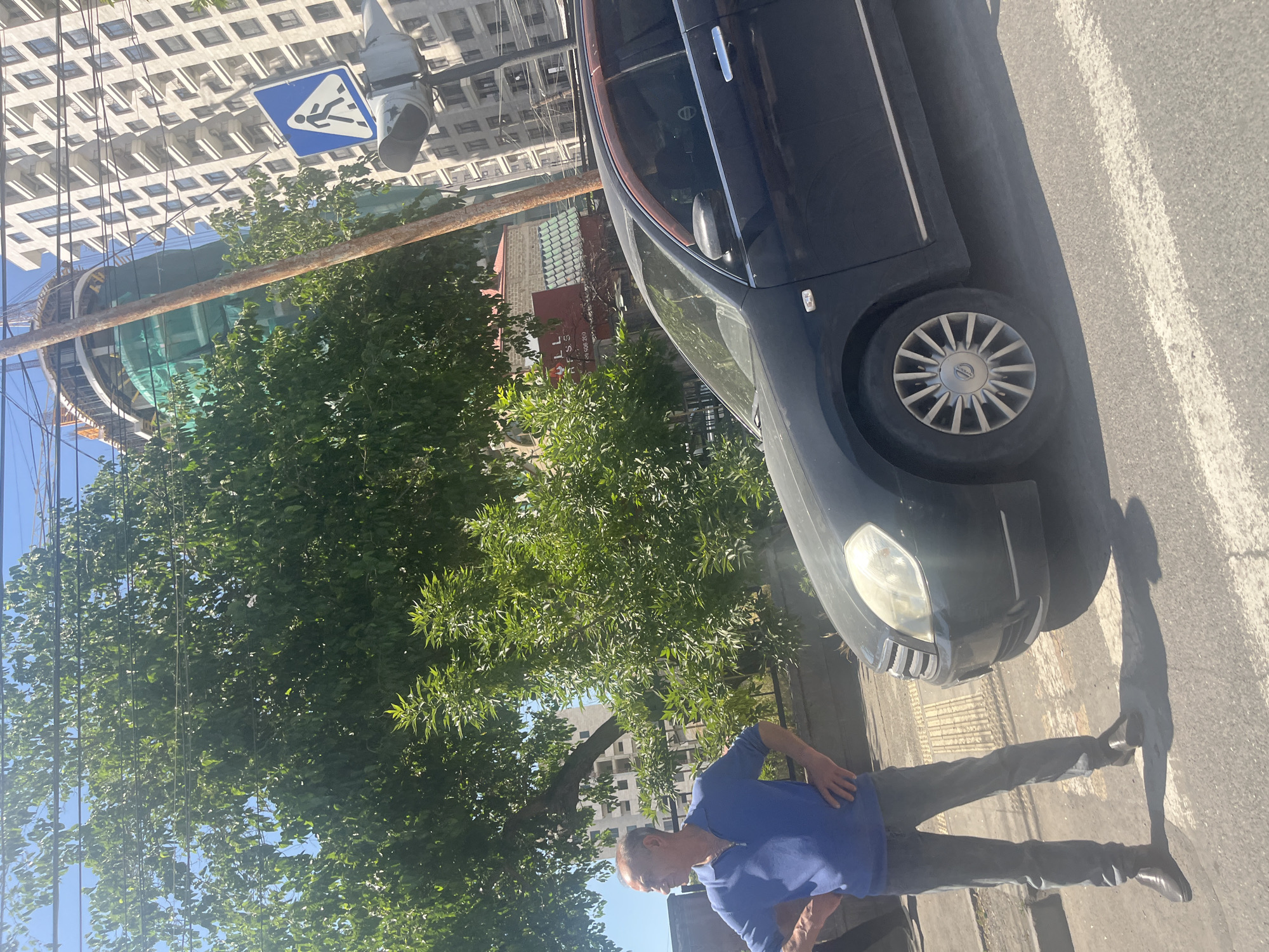 This image depicts a sunny street scene in an urban environment. In the foreground, a man wearing a blue polo shirt and dark trousers stands on a sidewalk, facing away from the camera with his right hand resting on his hip. A dark-colored car, possibly a sedan or hatchback, is parked next to him, with its front end pointing towards the right side of the frame and its front wheel partially on the sidewalk. The car features multi-spoke silver wheels.
Behind the man and the car, vibrant green trees dominate the mid-ground. In the background, several buildings are visible through and above the trees. To the upper left, a tall, modern building with a grid of square windows stands prominently. Below it and further in the distance, a large, circular, glass-and-metal structure appears to be under construction, partially covered by green netting, with construction cranes visible nearby. A rusty metal pole runs diagonally across the middle of the frame.
A blue triangular road sign, featuring a white walking figure (a pedestrian crossing sign), is attached to a pole in the upper left corner, near the modern building. The asphalt road in the foreground shows white painted markings. The bright sunlight indicates it is daytime, likely late morning or early afternoon, with clear skies. On a building partially obscured by trees, some red text, possibly "LL FSS" and numbers like "MT 600-300," is faintly discernible.