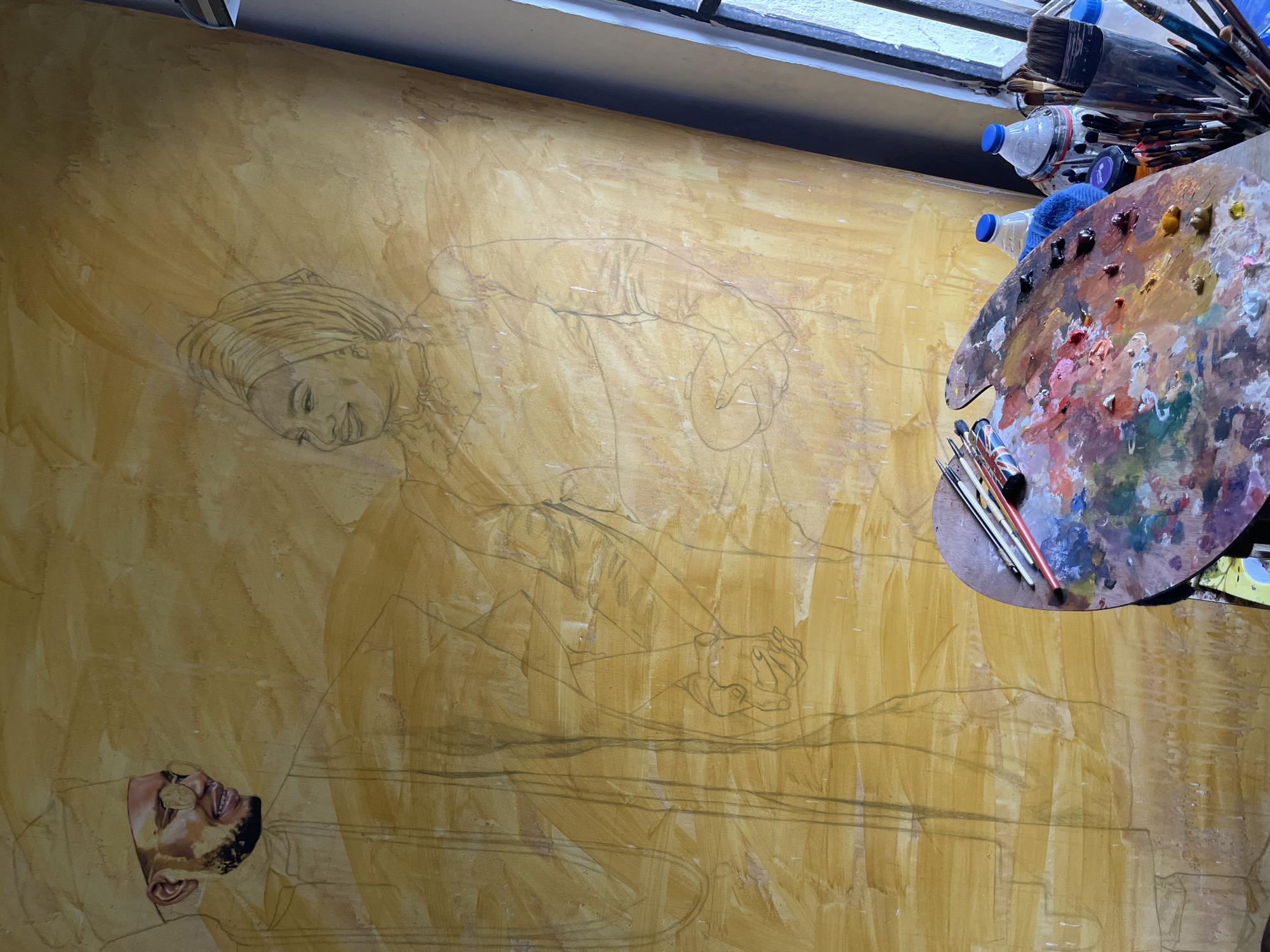 The image shows an artist's workspace, likely indoors in Ado, Nigeria. An unfinished painting covers the left side of the image; it features two figures: a woman with her head covered and a man in uniform. The figures are drawn with pencil on a yellow-toned background. To the right of the painting sits an artist's palette covered in oil paints, along with paintbrushes and water bottles. Above the palette, more brushes are stored on a shelf. The environment suggests an art studio or workshop.