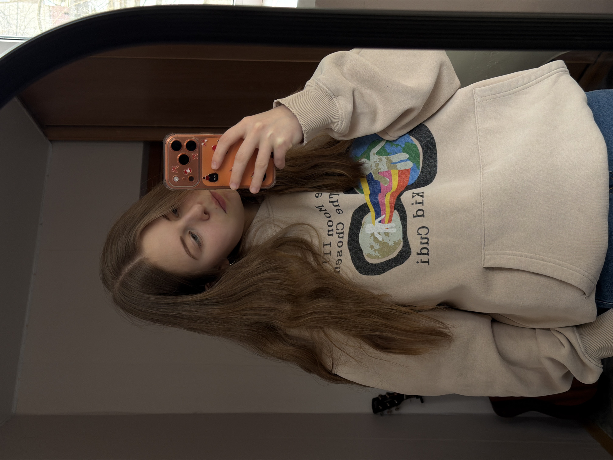 A young woman is taking a mirror selfie indoors. She is holding an orange iPhone in her right hand. She has long brown hair and is wearing a beige hoodie with an image of a world with colorful beams. She is also wearing blue jeans. In the mirror's reflection, the background shows what appears to be a wooden cabinet and a section of a room with a guitar. The location of the scene in the city of Perm, Russia cannot be determined based on the image.
