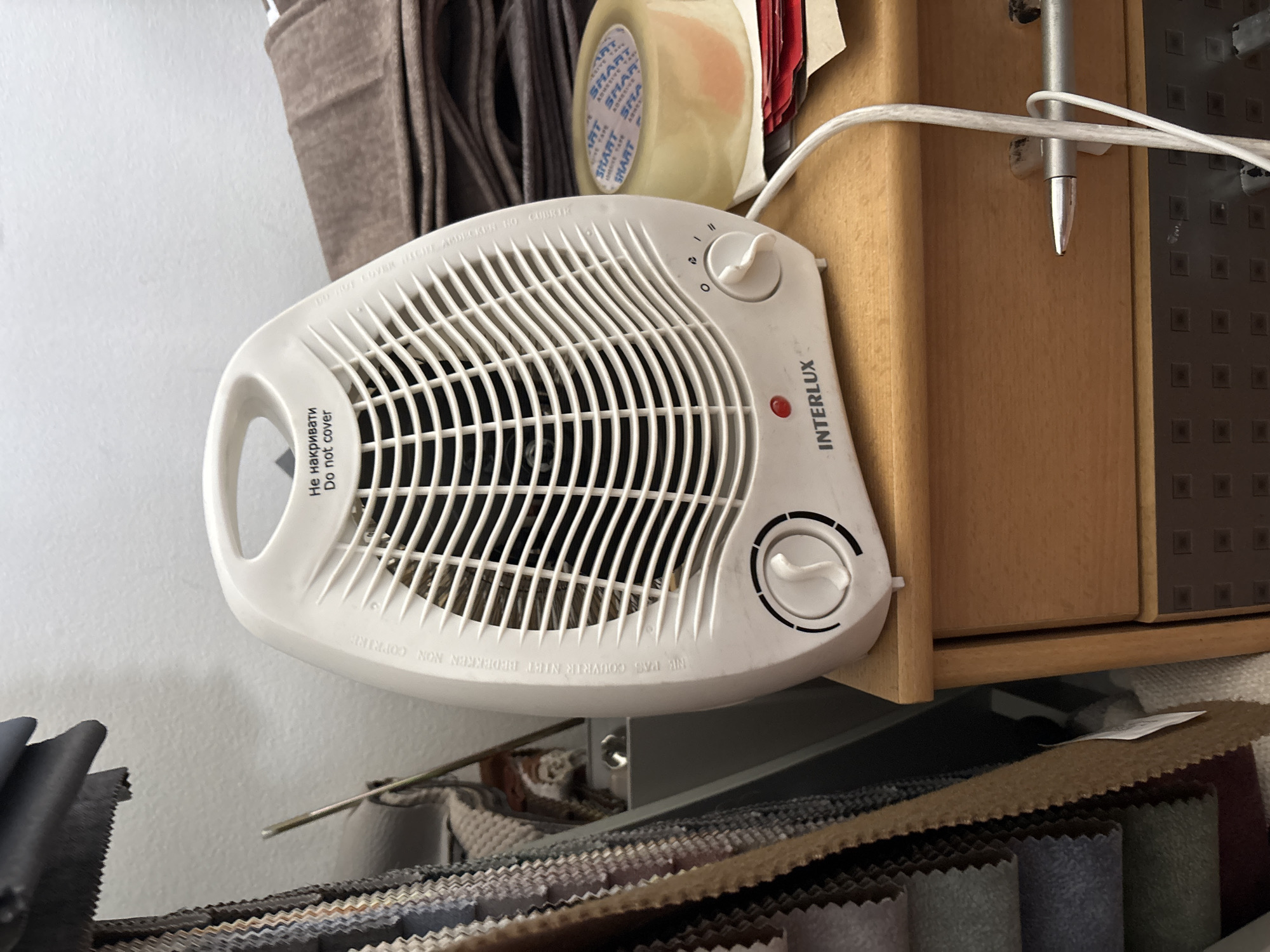 In Zaporizhzhia, Ukraine, a white electric heater with the brand name "INTERLUX" is placed on a wooden drawer. The heater has a warning label that reads "Не накривати Do not cover." To the right of the heater, a roll of clear adhesive tape is visible, partially obscuring some red paper items. A white electrical cord extends from the heater and is routed along the edge of the drawer, secured by a silver clip. Next to the clip, a silver pen rests horizontally on the drawer's surface. The background shows stacked fabric samples and a wall. The overall scene suggests a domestic or office environment where the heater is in use or ready for use.