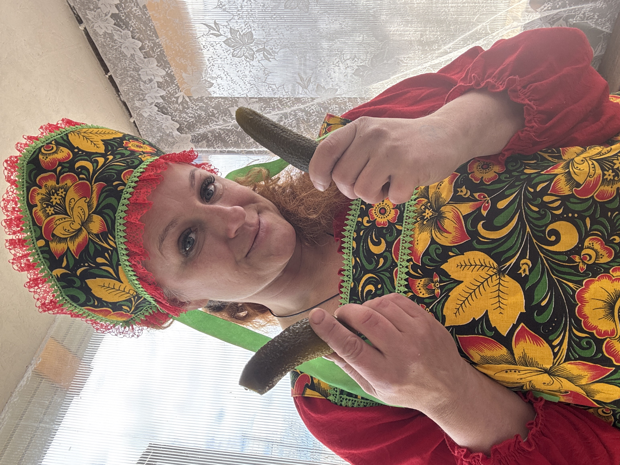 A woman in traditional Russian attire holds two pickles. She wears a floral headdress with a red lace trim, a red blouse with ruffled sleeves, and a floral print dress. She is looking directly at the camera with a slight smile. The background is a window with a floral lace curtain, likely inside a building. Based on the cultural attire, it can be inferred this scene is likely set in Russia, potentially in a location like Chaikovsky.