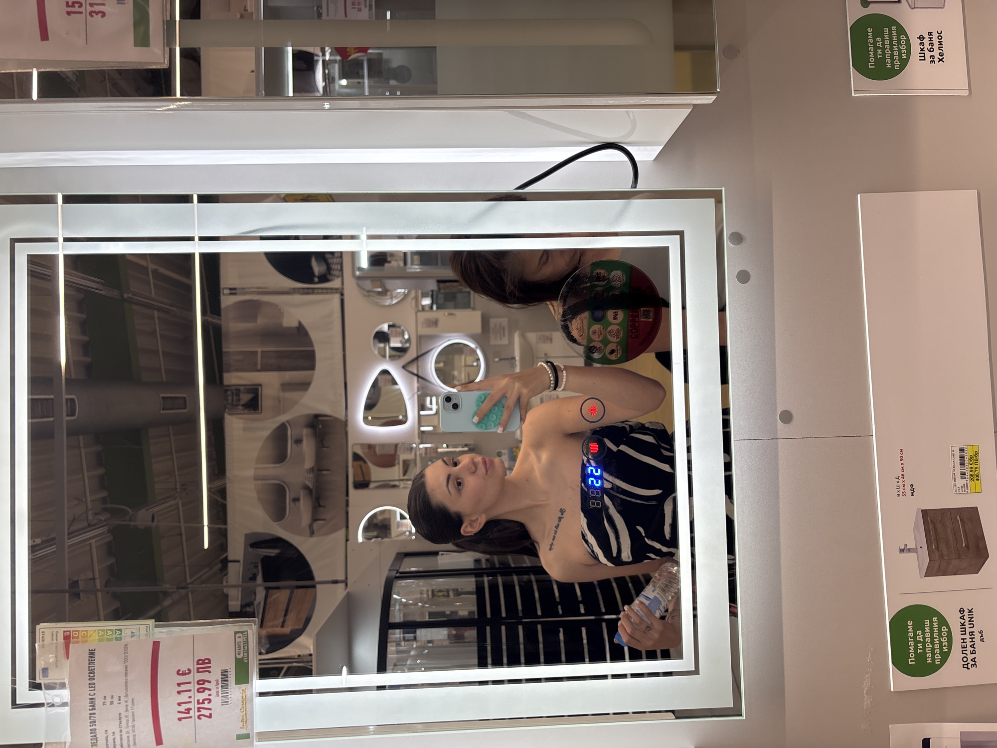 The image depicts a young adult female taking a selfie in a large, rectangular, LED-illuminated mirror within a retail store in Sofia, Bulgaria. The location is confirmed by the presence of Bulgarian Cyrillic text on product labels, the currency "ЛВ" (Bulgarian Lev), and the "Inter Ceramic" brand name on a price tag reflected in the mirror.
The primary subject, reflected in the mirror, has dark hair tied back in a ponytail and wears a black and white zebra-print strapless top. She holds a light blue smartphone with a distinct pop-it case in her right hand, pointed towards the mirror for the selfie. In her left hand, she holds a transparent plastic water bottle. A tattoo of text is visible on her left collarbone area. The smart mirror itself features a digital display showing "88:22" in blue, flanked by two circular touch controls: a red one with a wavy line icon and another with a sun-like icon.
To the right of the main subject's reflection, a second individual, likely female, is partially visible. Only her upper head and shoulder are seen, looking downwards, and she appears to be holding a circular item bearing the text "COPPER." A black electrical cable descends from the right side of the mirror, partially obscuring this second individual.
The background reflection within the mirror reveals various other mirrors of different shapes, bathroom sinks, and general store shelving, suggesting a showroom environment for home improvement or bathroom fixtures. In the immediate foreground, below the mirror's frame, product information is displayed on white signs with green accents. These signs promote "ДОЛЕН ШКАФ ЗА БАНЯ UNIK" (Lower cabinet for bathroom UNIK) and "ШКАФ за баня Хелиос" (Cabinet for bathroom Helios), providing details such as dimensions (e.g., 55 cm x 46 cm x 50 cm), material (MDF), and prices in both Euros (€141.11) and Bulgarian Levs (275.99 лв). The overall store environment features light-colored walls and overhead industrial lighting.