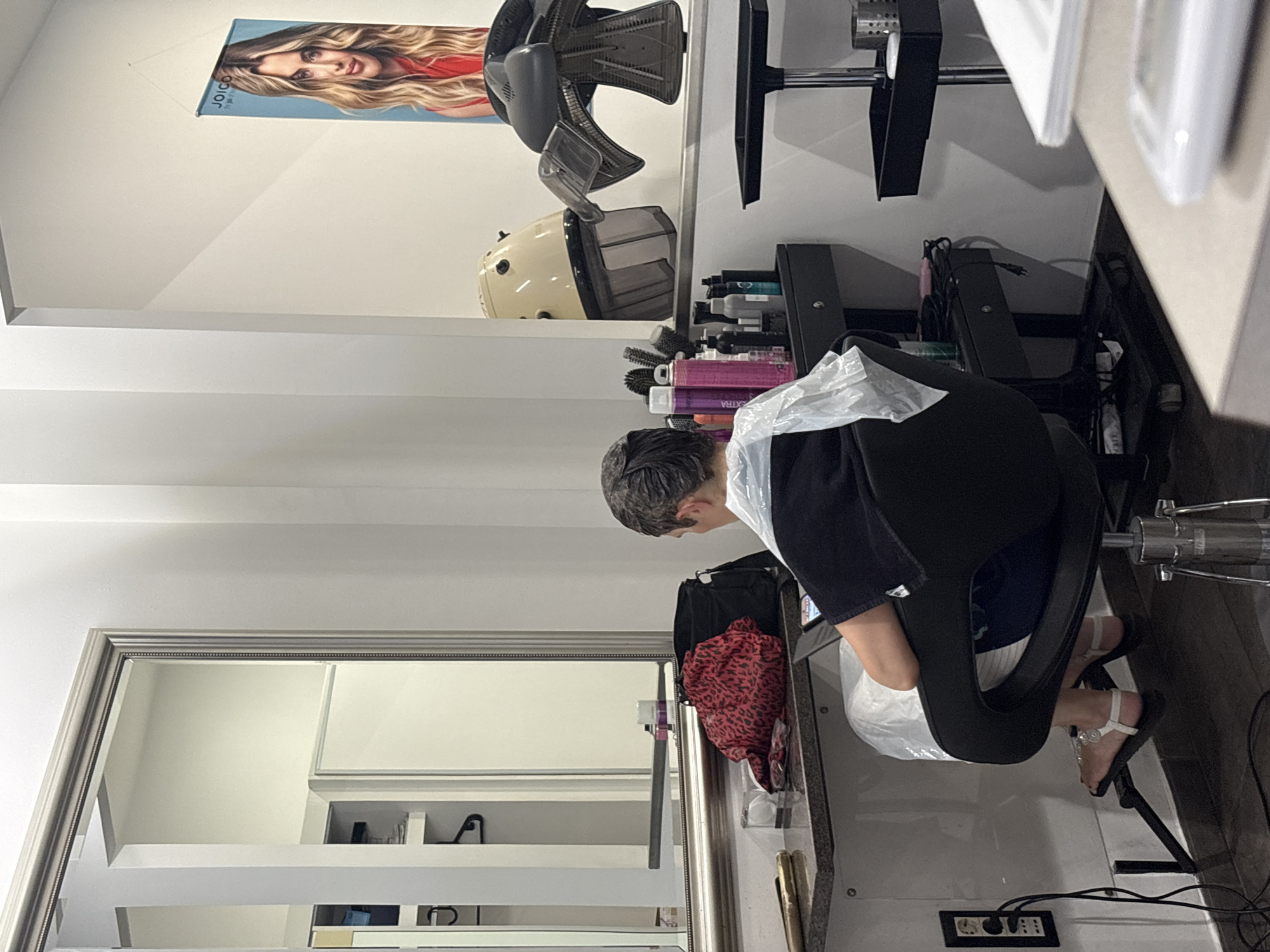 The image depicts the interior of a hair salon, potentially located in Rome, Italy, as suggested by the request. A person with short, graying hair sits facing away from the camera in a black salon chair, covered with a clear plastic cape. Their hair appears to be treated with a product. They are holding a mobile device, possibly a tablet, and are looking at it.
In the background, a large mirror reflects a portion of the salon, including shelves and what appears to be a closet. To the right of the person, a black trolley holds various hair care products, including several aerosol cans and bottles of shampoo or conditioner. A beige hooded hair dryer is visible, as is a black overhead hair dryer. A poster featuring a blonde woman with the "JOICO" logo hangs on the wall.
On the counter in front of the person, a red and black animal print scarf or fabric is draped over a black bag. A pair of sunglasses rests on the counter as well. Electrical outlets and cords are visible on the wall and floor, indicating the presence of salon equipment. The flooring appears to be tiled. The overall impression is a functional, possibly a bit cluttered, working salon environment.