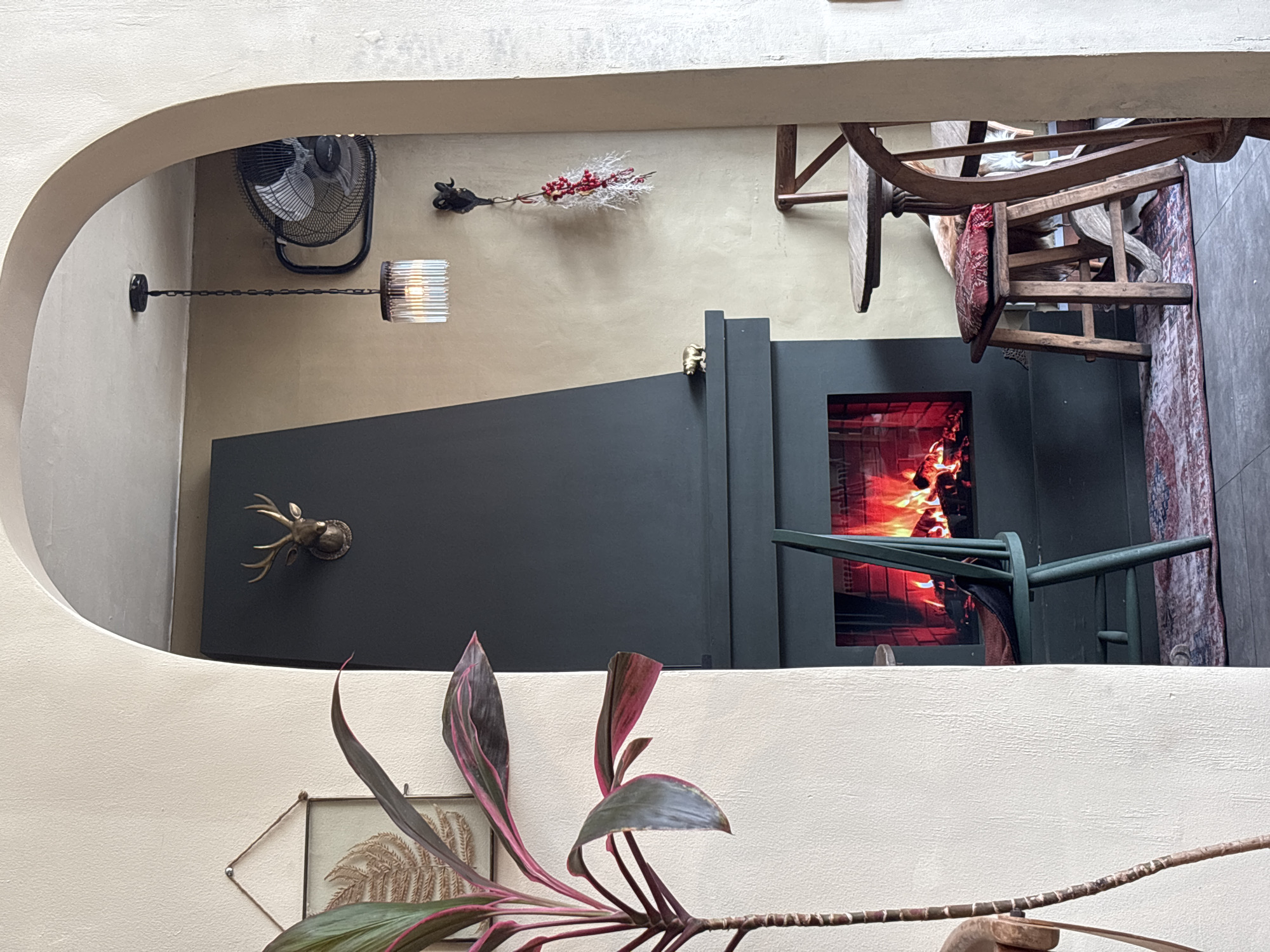 A high-angle indoor shot, taken through an arched opening in a wall, reveals a section of a room. The focal point is a dark, possibly black, enclosed fireplace with a rectangular opening displaying flames. To the left of the fireplace, attached to the wall, is a metallic deer head. A wall-mounted lamp with a cylindrical, ribbed shade hangs near a black fan. Above the fireplace is a decorative twig with red berries, and part of a wooden chair with a patterned cushion is visible. In the foreground, a plant with reddish-pink leaves and a framed botanical print are positioned near the base of the arched opening.
The location is identified as Kuta Selatan, Indonesia.