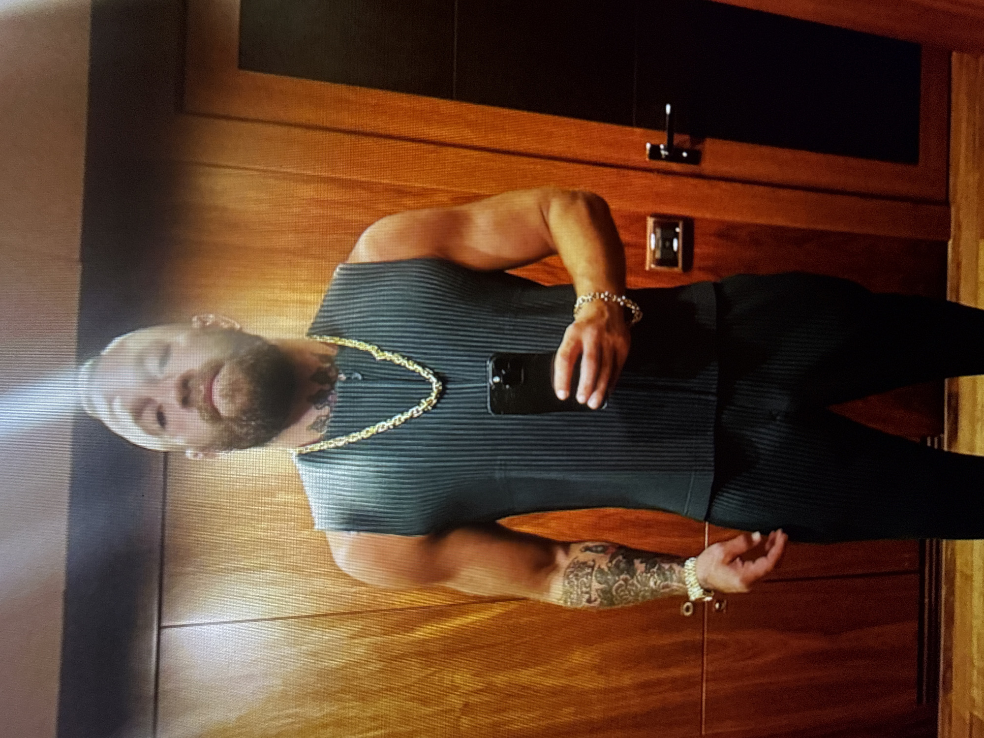 A man is standing in a room with wood-paneled walls, holding a black smartphone in his right hand. He is wearing a sleeveless, dark gray, pleated top, black pants, a thick gold chain necklace, and a silver bracelet on his left wrist. He has tattoos on his left arm and neck. The room appears to be a hallway or a room with a door on the right side. There is a light fixture on the wall to the left of the door, and a door handle on the door itself. The man is looking directly at the camera. The scene appears to be indoors, and there is no indication of the city of Tbilisi, Georgia.