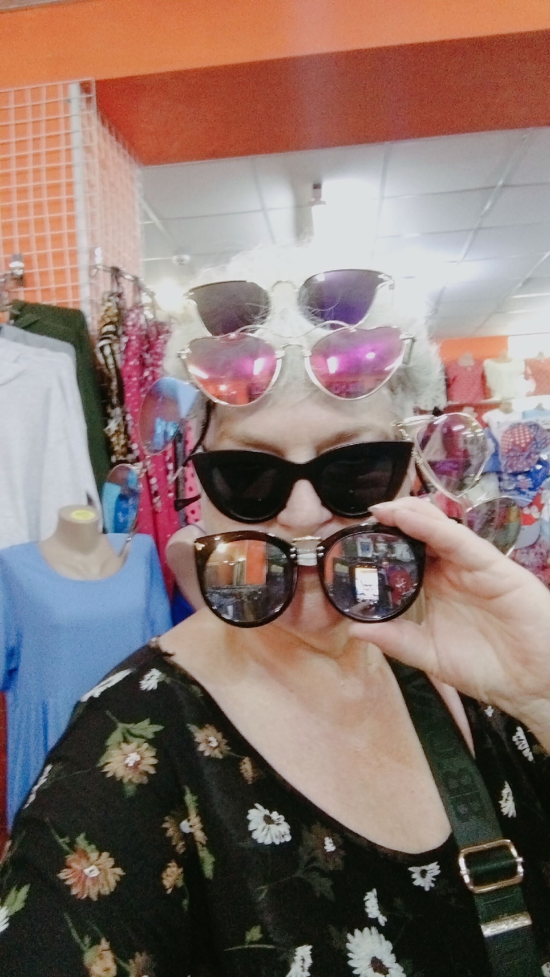 A woman with white hair and glasses is posing for a selfie in what appears to be a store in Horishni Plavni, Ukraine. She is wearing a black floral-patterned top and has multiple pairs of sunglasses stacked on her head, with one pair held up to her face. Behind her, clothing racks and displays of merchandise are visible. The store has orange and white shelving and white ceiling tiles.