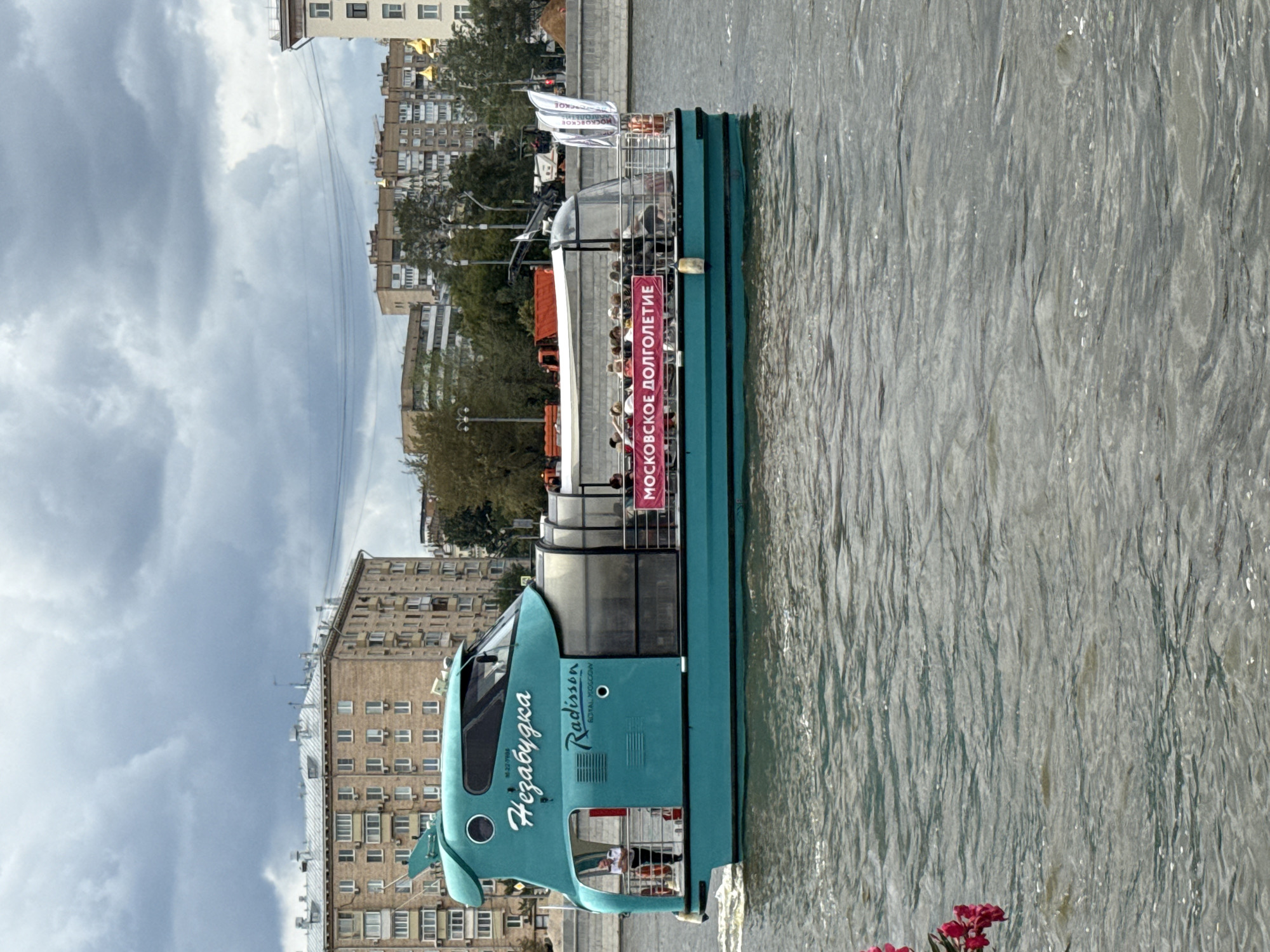 The image shows a teal-colored boat named "Незабудка" (Nezabudka) on a body of water, likely a river, in Moscow, Russia. The boat has a modern design with a large glass-enclosed passenger cabin. There are people visible inside the cabin, seated and appearing to be on a tour. A banner on the side of the boat reads "МОСКОВСКОЕ ДОЛГОЛЕТИЕ" (Moscow Longevity). On the front of the boat, the name "Radisson" is visible, suggesting it is operated by Radisson Royal, Moscow, which offers river cruises.
In the background, there are several multi-story residential buildings with a Soviet-era architectural style. Trees and some construction equipment are also visible along the embankment. The sky is overcast with dramatic clouds. The water is choppy, indicating the boat is in motion. The overall scene depicts a typical summer day in Moscow with a popular tourist activity.