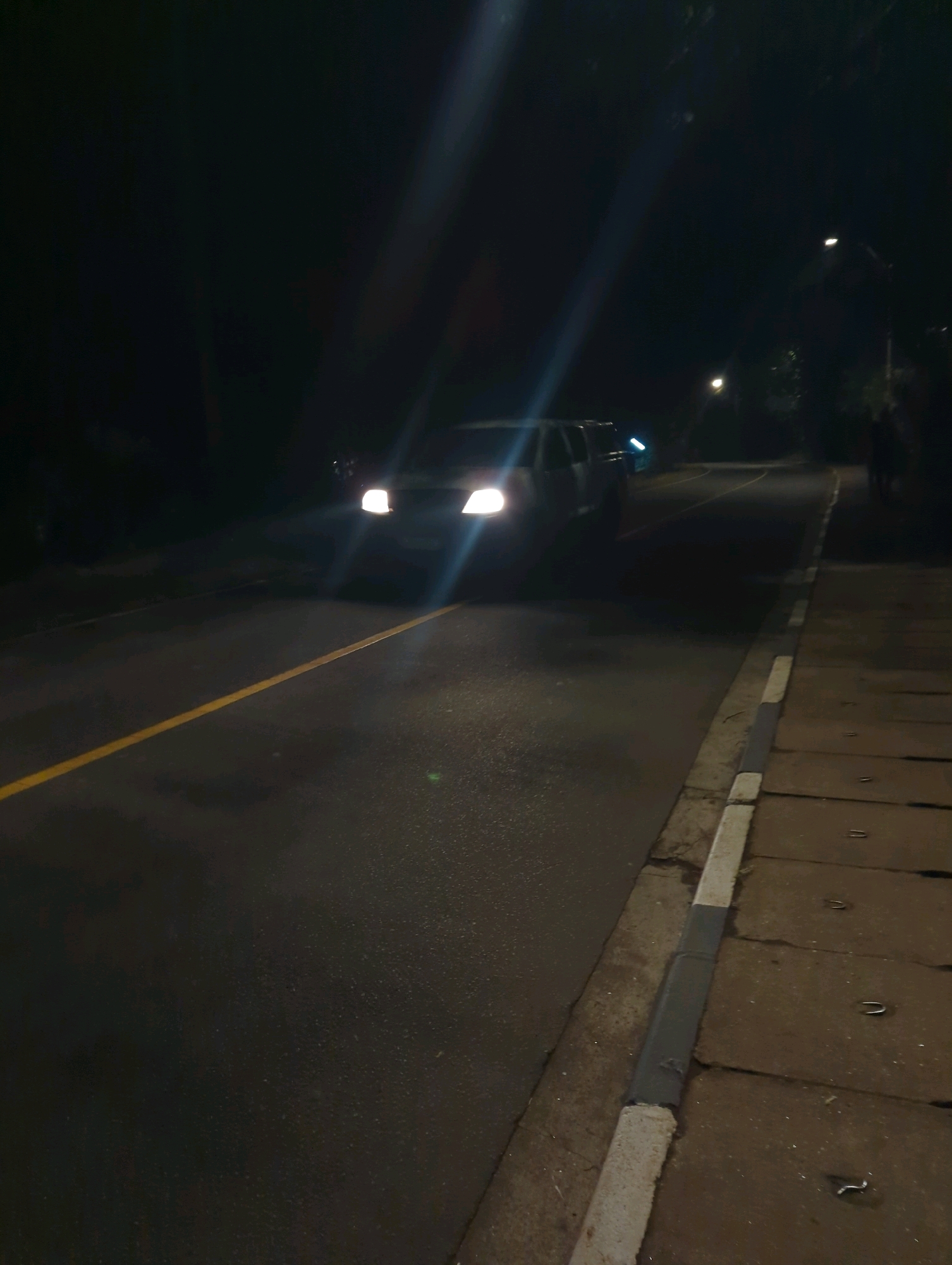 An image taken at night on a road in Musanze District, Rwanda. A white van with its headlights on is driving on the road. The road has a yellow line marking the center. To the right of the road, there is a sidewalk with concrete slabs. There are streetlights visible in the background, and trees line the side of the road. The image appears to be taken from a slightly elevated position, looking down the road.