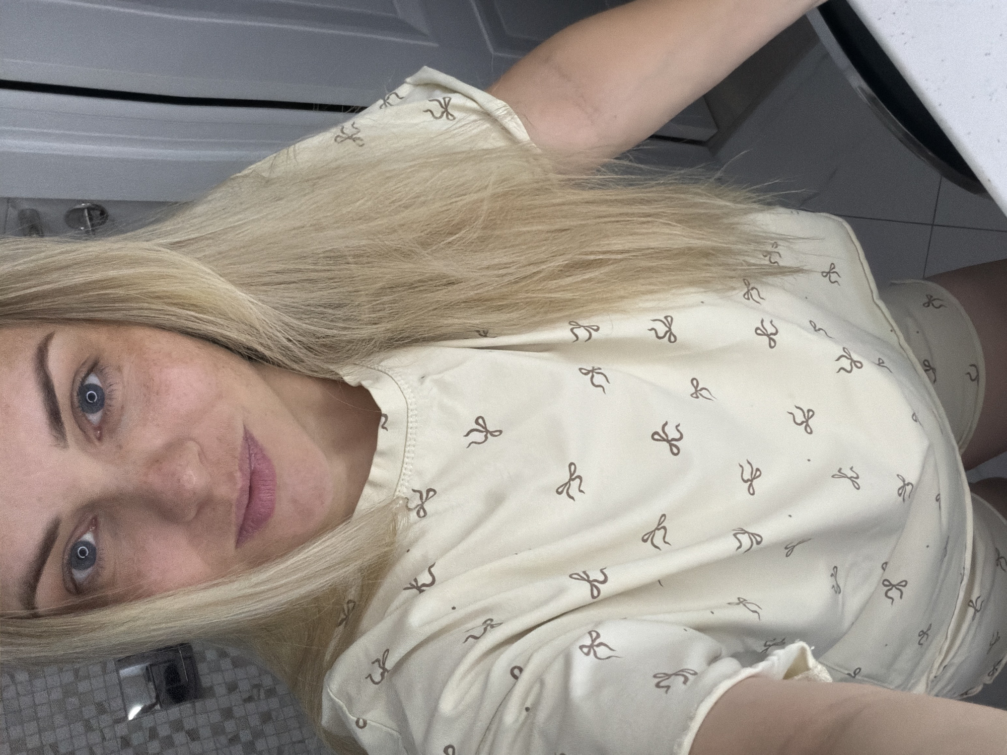 A young woman with blonde hair and blue eyes is taking a selfie. She is wearing a light-colored pajama set with a repeating bow pattern. Her head is tilted, and her gaze is directed at the camera. Her skin has a natural appearance with freckles visible. She is in an indoor setting, with tiled flooring and what appears to be a bathroom counter and cabinetry visible in the background. The lighting suggests it might be daytime, though no direct sunlight is evident. The overall mood is casual and relaxed. No text is visible in the image.