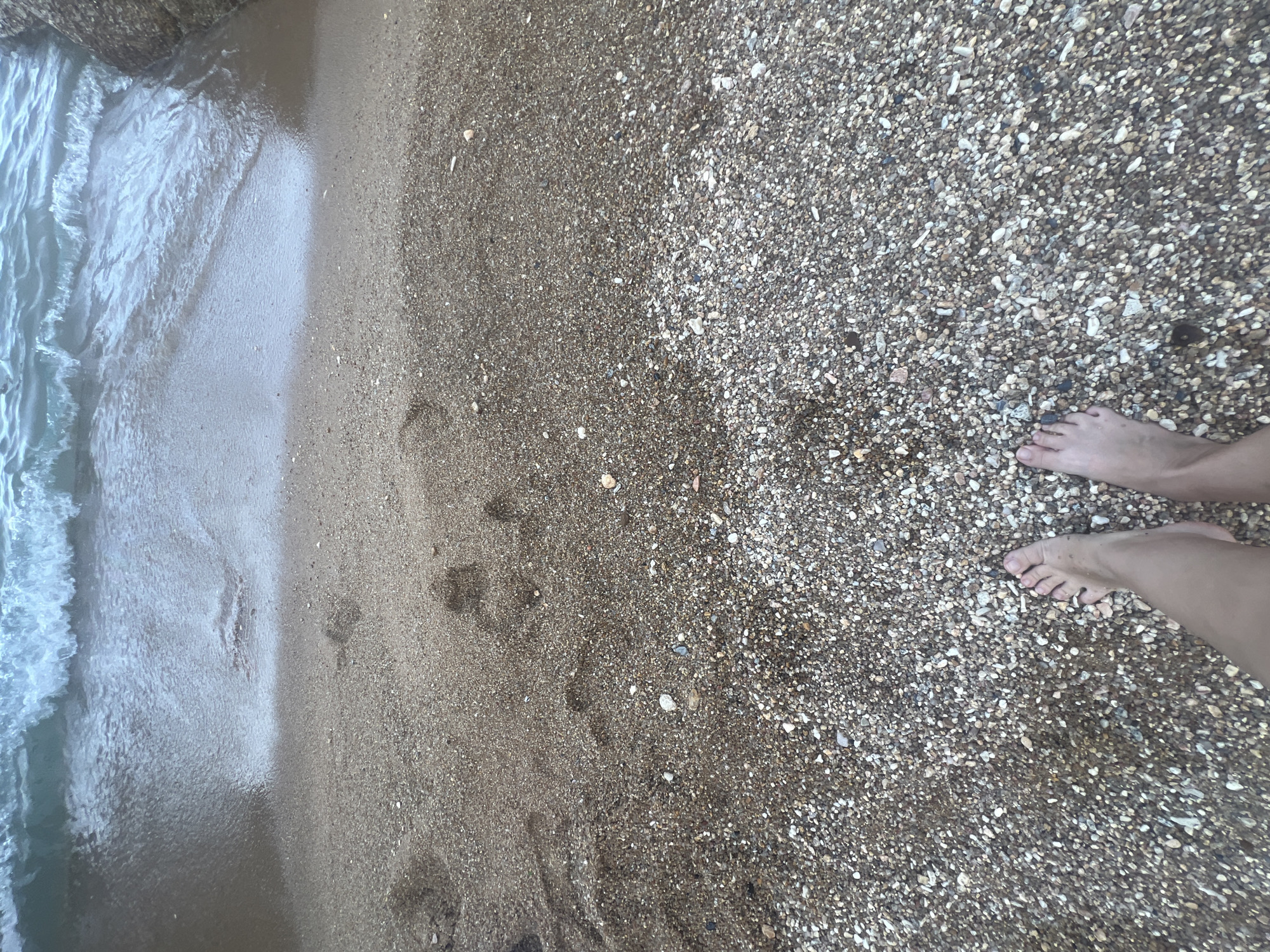 The image shows a first-person perspective of a person standing on a beach in Ko Samui District, Thailand. The beach consists of sand and small pebbles. The sea is visible at the top of the image with gentle waves approaching the shore. There are footprints visible in the wet sand near the water's edge. The person's bare feet are in the foreground, standing on the pebble and sand mixture. The lighting suggests it is daytime, possibly overcast or during the golden hour.