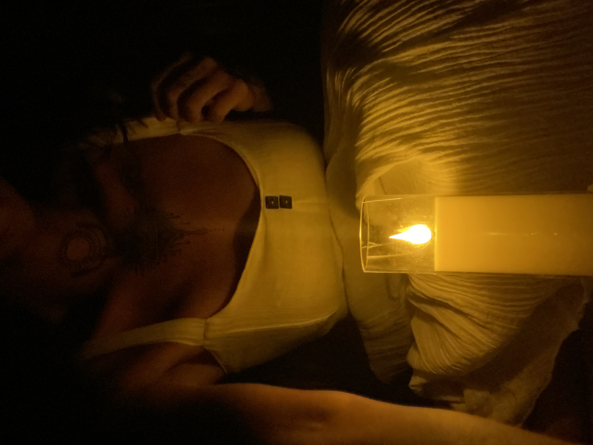 In a dimly lit room, a person sits wearing a white garment. A lit candle in a glass holder is positioned in the foreground, casting a warm glow on the textured fabric of the garment. The person's upper body is visible, with tattoos on their chest and neck. Their hands are partially visible, one near their hair and the other resting on their lap. The background is dark, with no discernible details of the surroundings. There is no information to suggest this scene is located in Mui Ne, Vietnam.