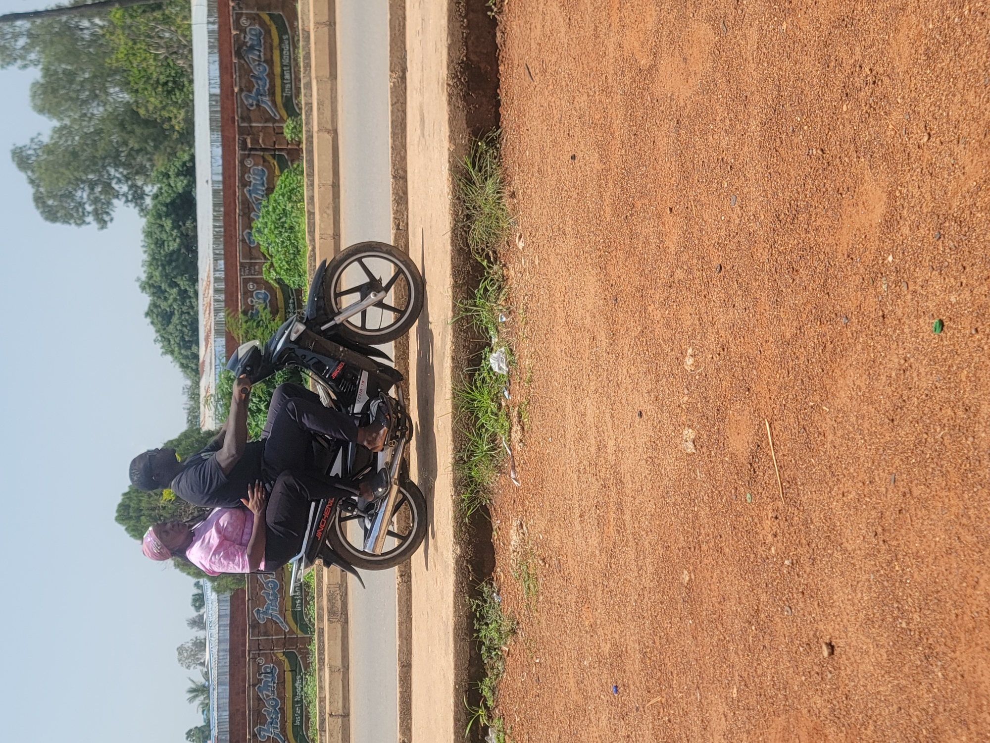 In Barakin Lerawi, Nigeria, a motorbike carries three people down a dirt road. The rider, a man wearing a black t-shirt and dark pants, is at the front. Behind him, a woman in a pink t-shirt and headscarf sits with her arm around his waist. A third person, also a man in dark clothing, is perched behind the woman. The motorbike is black with silver accents, and its wheels are clearly visible. The road is unpaved and appears to be made of reddish-brown dirt. To the left of the road, there's a paved walkway or sidewalk with a low brick wall. Behind the wall, a row of buildings with graffiti-style advertisements can be seen, with lush green trees and foliage visible in the background. The sky is bright and clear, suggesting a sunny day. The overall scene depicts everyday transportation in a rural or semi-urban Nigerian setting. The people on the motorbike appear to be casually dressed, and their posture suggests they are comfortable with each other. The advertisements on the wall seem to be for food products, with the words "Indo Mia" and "Instant Noodles" visible.