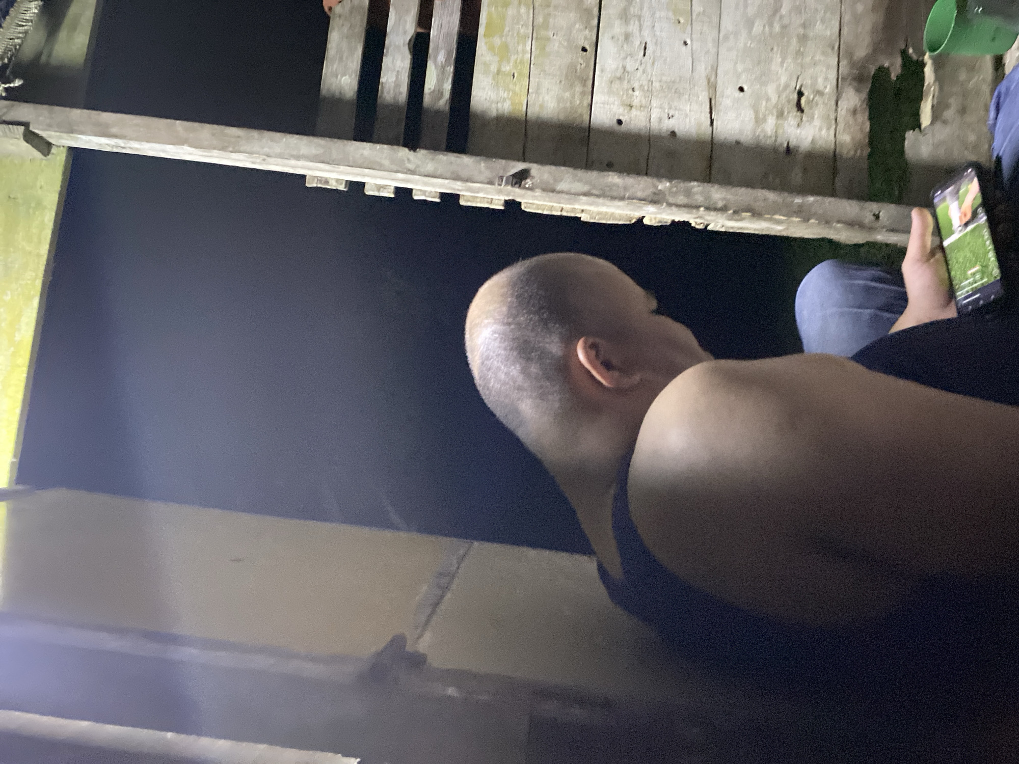 A man with a shaved head is sitting on the ground, looking at a smartphone. He is wearing a dark blue tank top and blue jeans. The background is dark, with some wooden structures visible. The scene appears to be outdoors at night. The location is identified as Lamtui, Indonesia, although this information is not visually verifiable from the image itself.