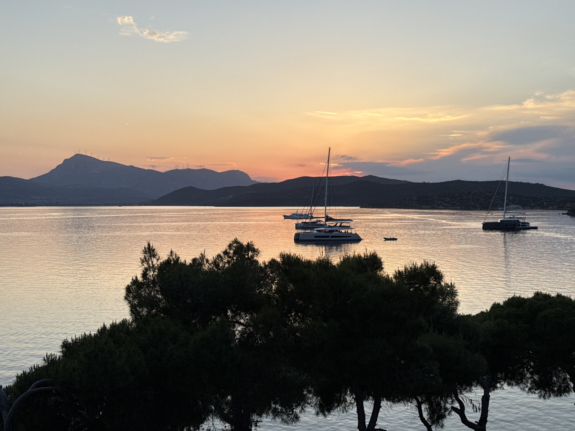 The image depicts a sunset scene over the sea near Poros, Greece. In the foreground, dark green trees partially obscure the view. The middle ground features the calm sea with several sailboats and a motor yacht. The background consists of mountainous land, with the setting sun casting a warm, orange glow across the sky and water. A cluster of wind turbines can be seen atop the highest mountain.