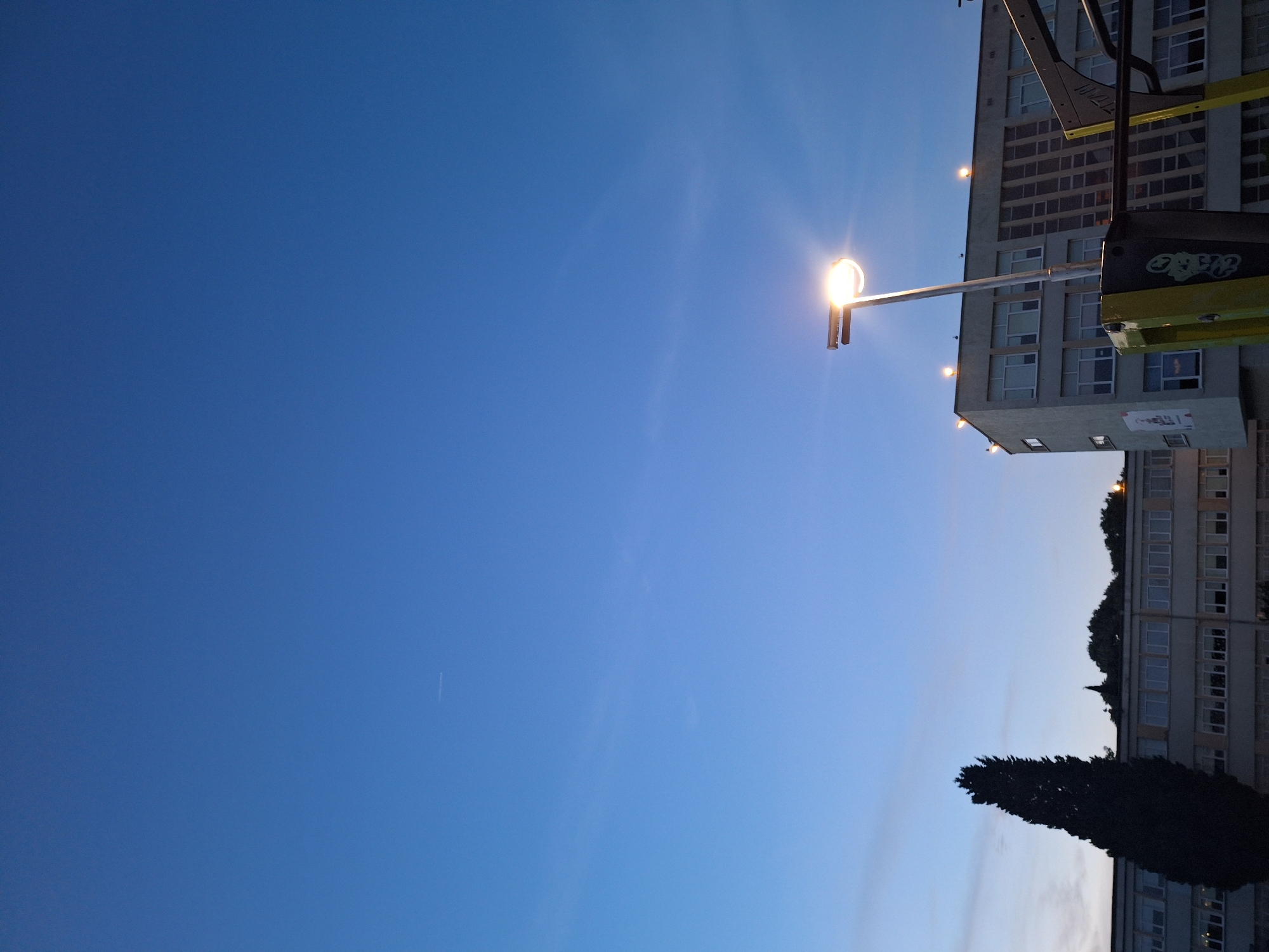 The image depicts a wide shot of a building's facade and the sky. The sky is a gradient of blue, with a faint contrail visible near the top left. A street lamp is illuminated in the lower right quadrant of the image, casting a bright light. A tall, dark cypress tree stands to the left of the building, silhouetted against the sky. The building itself appears to be a school or office building, with multiple windows. The overall scene suggests it is dusk or dawn. The image is likely taken in Varna, Bulgaria, based on the presence of the street lamp, typical architecture, and cypress tree, which are common in the region.