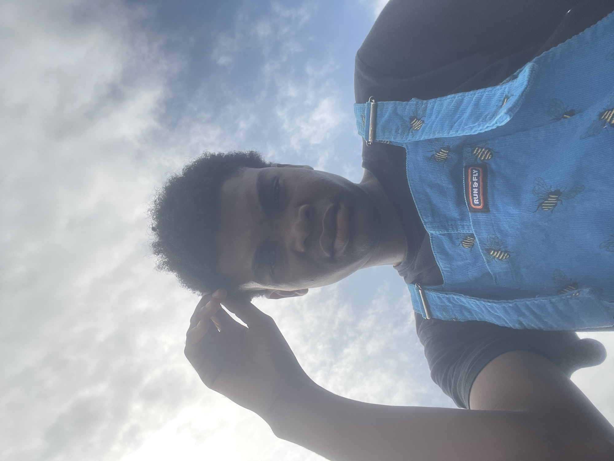 A young Black person, with dark, curly hair and medium-brown skin, is shown from a low angle, looking up at the camera. They are wearing a blue corduroy overall with a bee pattern and a black t-shirt underneath. A patch on the overalls reads "RUN & FLY". The person's left hand is raised to their forehead. The background is a sky with clouds. The image appears to have been taken outdoors on a bright, overcast day. There is no indication of the location being Ilado, Nigeria.