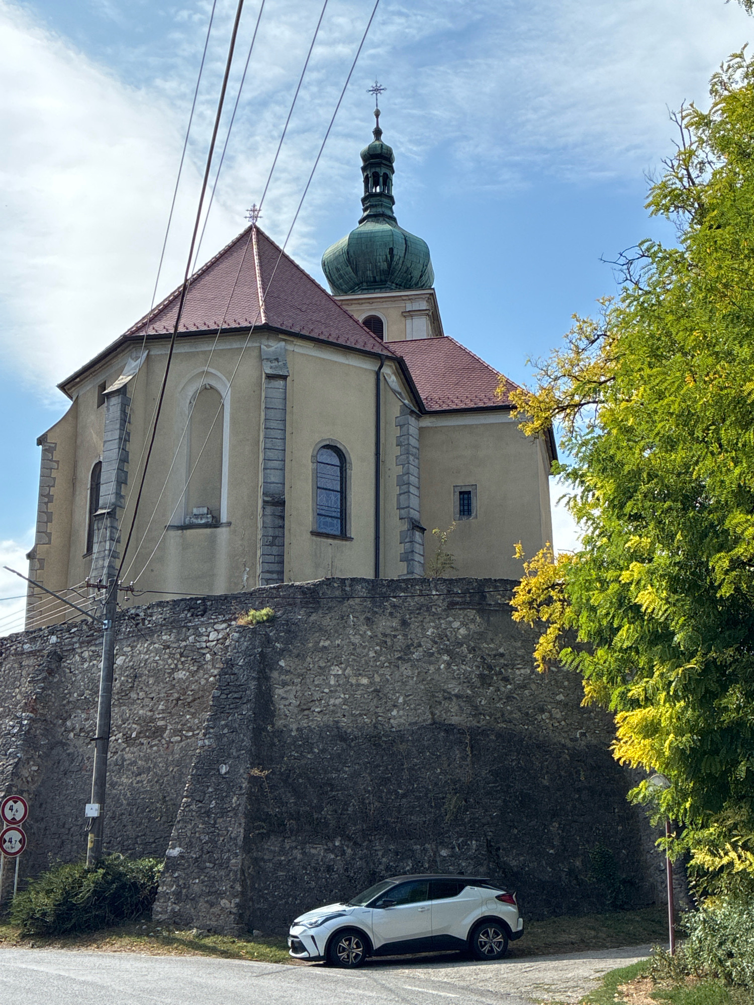 The image shows a stone wall with a church built on top of it, in the town of Čachtice, Slovakia. A white car is parked on the side of the road in front of the wall. The church has a red tiled roof and a green dome. A large tree with green and yellow leaves is on the right side of the image. The sky is blue with some clouds. There are power lines running across the top of the image.
