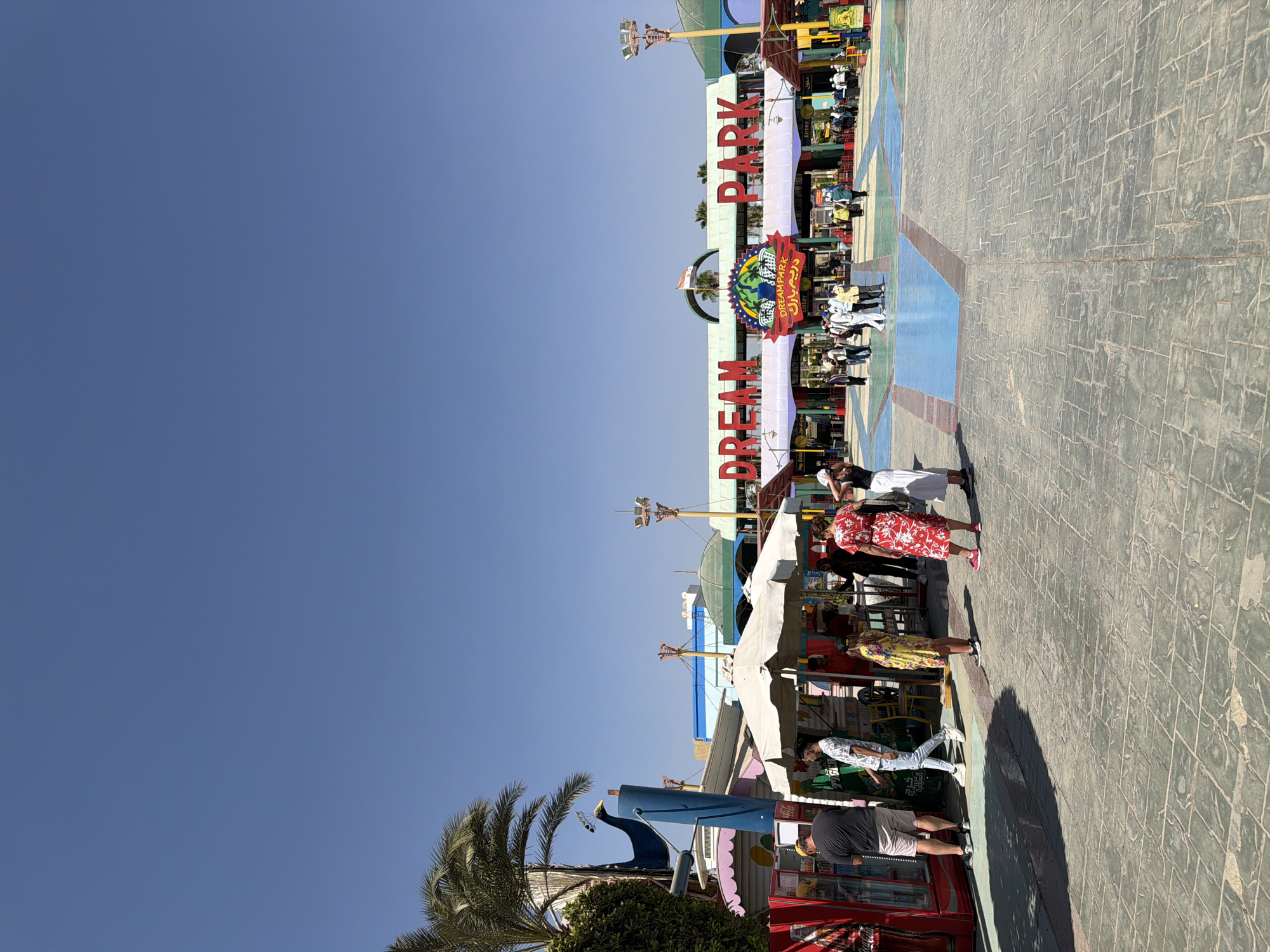 The image shows the entrance to Dream Park in Sheikh Zayed City, Egypt. The park's sign, "DREAM PARK," is prominently displayed in large red letters above the entrance. The area in front of the entrance is paved with stone tiles and has some colorful painted sections.
Several people are visible in the scene. In the foreground on the left, a man wearing a black t-shirt and shorts is standing near a red Coca-Cola branded vending machine. To his right, a man in a white patterned t-shirt and jeans is walking. Further back, a woman in a yellow floral dress stands near a white umbrella. More people are gathered near the park entrance, some entering and others standing around.
To the left of the entrance, there are several stalls or kiosks with colorful facades, one of which has a large blue peacock statue on top. Palm trees are also visible in the background, along with various amusement park structures and rides. The sky is clear and blue, indicating a sunny day.