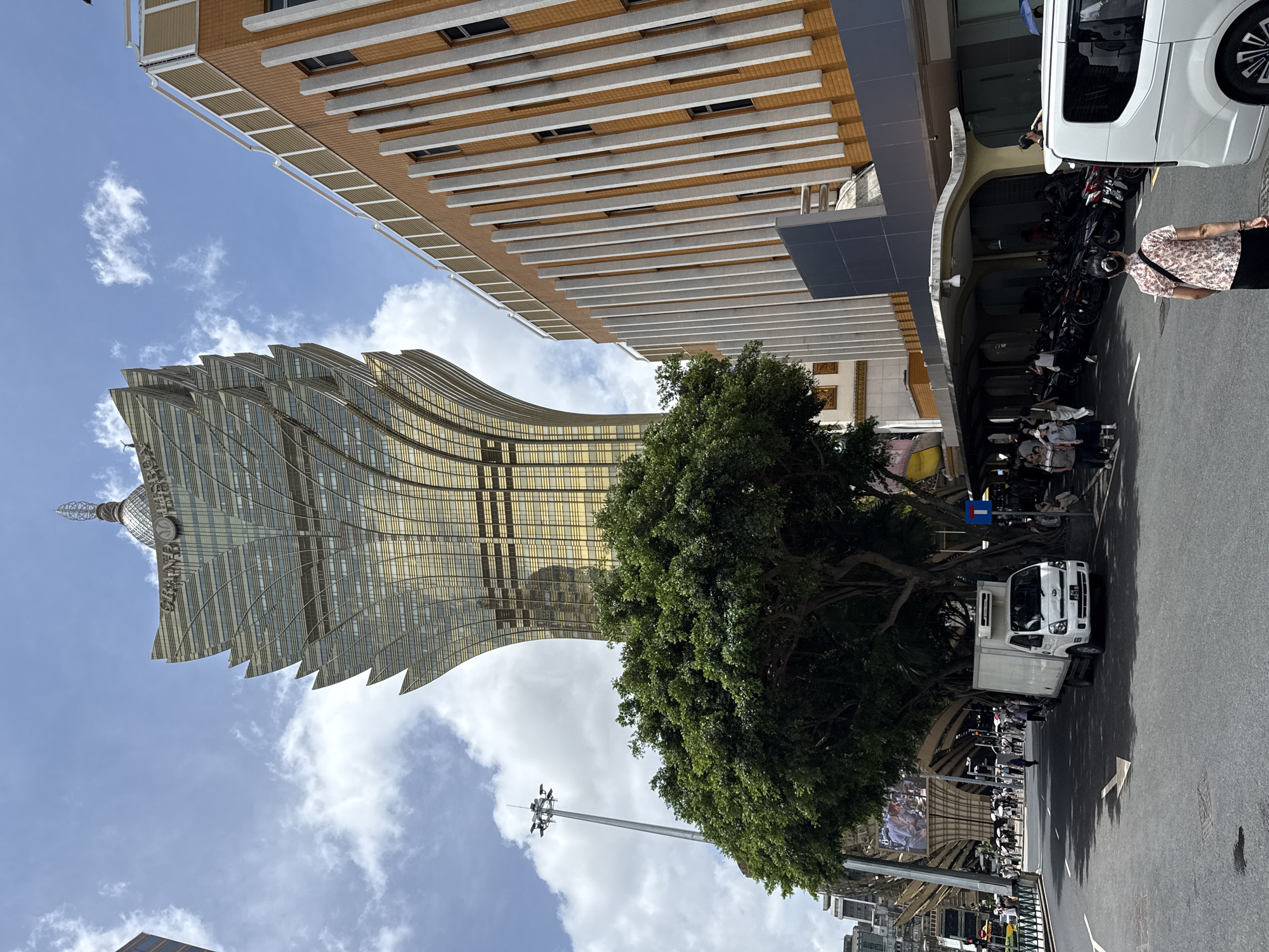 The image captures a street scene in Macau, China, with a prominent view of the Grand Lisboa Palace Resort Macau. The iconic golden, lotus-shaped skyscraper dominates the background, its distinctive architecture reaching towards a partly cloudy sky. In the midground, a large, lush green tree provides shade over a street and sidewalk. A white delivery truck is parked on the right side of the road, in front of the tree. To the right of the tree, a modern orange and white building with vertical striped accents is visible. The foreground shows a street with a white arrow painted on the asphalt, indicating a lane direction. Several people are seen walking on the sidewalk and along the street, some of whom are gathered near parked motorcycles. A white van is parked on the far right of the image, partially obscuring the view of the sidewalk. The overall atmosphere suggests a busy urban environment on a bright, sunny day.