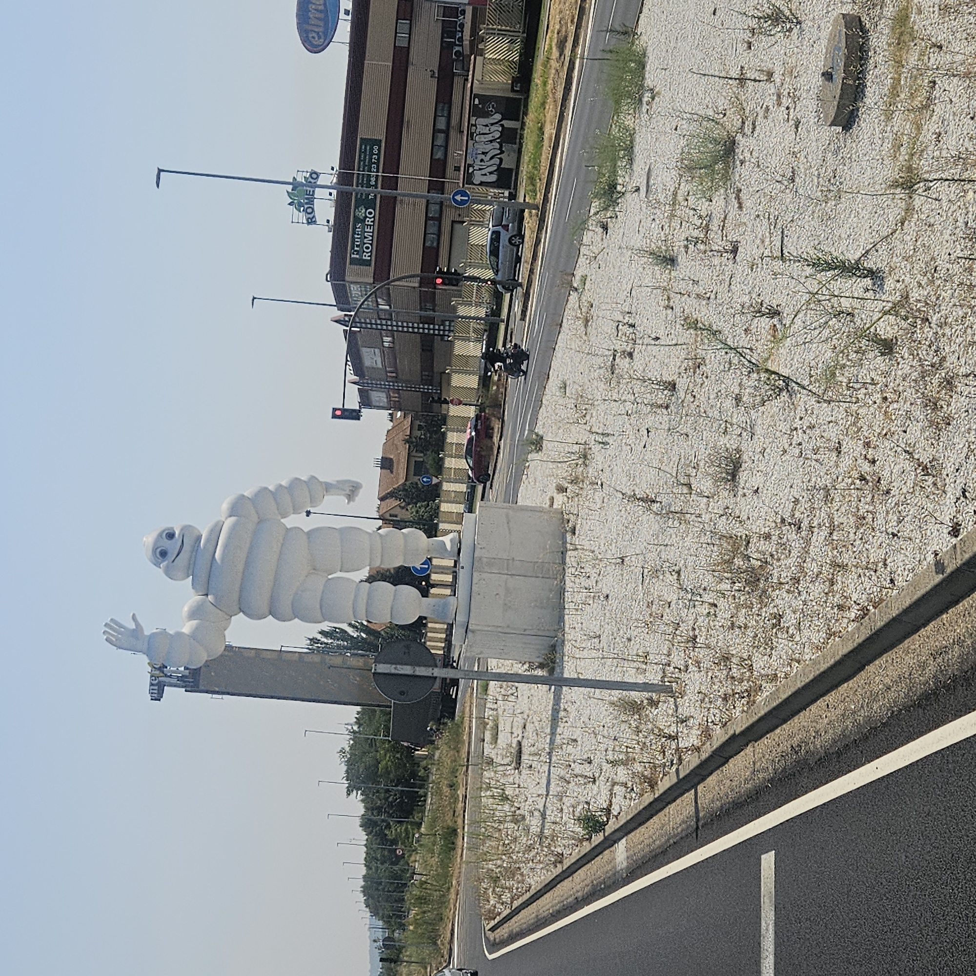 The image depicts a scene in Valladolid, Spain, featuring a large, white, inflated statue of the Michelin Man standing prominently on a concrete pedestal. The statue is on the left side of the image, its right arm raised in a wave. Behind the Michelin Man, a tall, slender tower with the Michelin logo at its top is visible.
To the right of the Michelin Man, a road runs horizontally across the frame. Several vehicles are present on the road, including a silver minivan, a red sports car, and a motorcycle ridden by a person wearing a helmet and black clothing. A traffic light shows a red signal. Beyond the road, a commercial building with striped brown and beige facades is visible, bearing signs for "FRUTAS ROMERO" and "elme". A fence with a grid pattern separates the building from the road.
The foreground of the image consists of a patch of ground covered in white gravel and sparse, dry vegetation. A concrete curb lines the edge of the road. The sky is clear and blue. The overall impression is an outdoor commercial or industrial area, possibly near a transportation hub, given the road and vehicles. The presence of the Michelin Man statue suggests a connection to the tire company, possibly a dealership or a historical landmark.