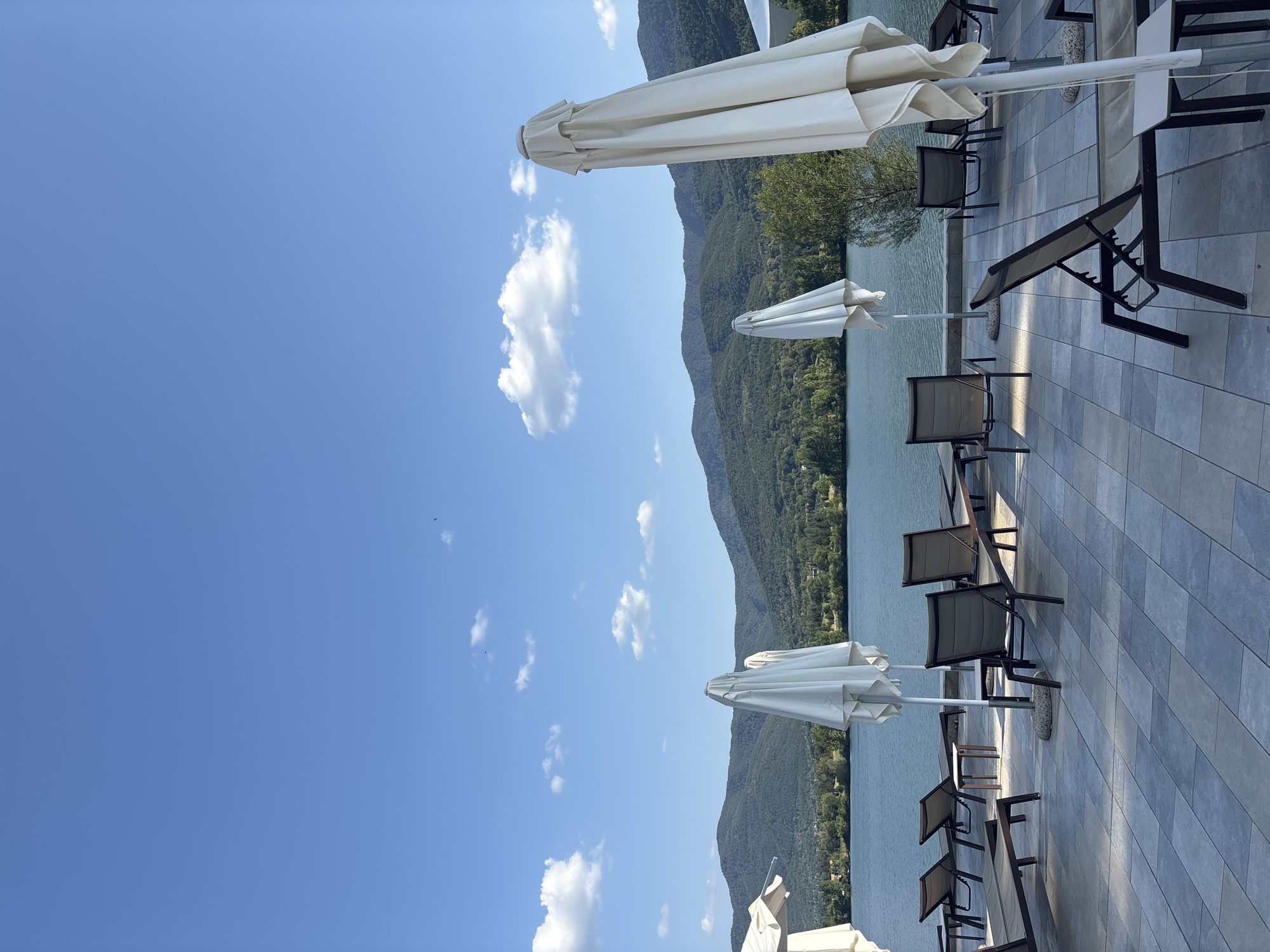 An exterior, eye-level shot captures a tranquil scene at a lakeside resort in Razliv, Bulgaria. The foreground is dominated by a paved patio with several empty lounge chairs and closed white umbrellas, suggesting a calm, possibly late afternoon setting. Beyond the patio, a serene blue lake stretches out, bordered by lush, tree-covered hills that form a backdrop. The sky above is a clear, vibrant blue with scattered white clouds. The overall impression is one of peaceful relaxation in a natural, scenic environment.