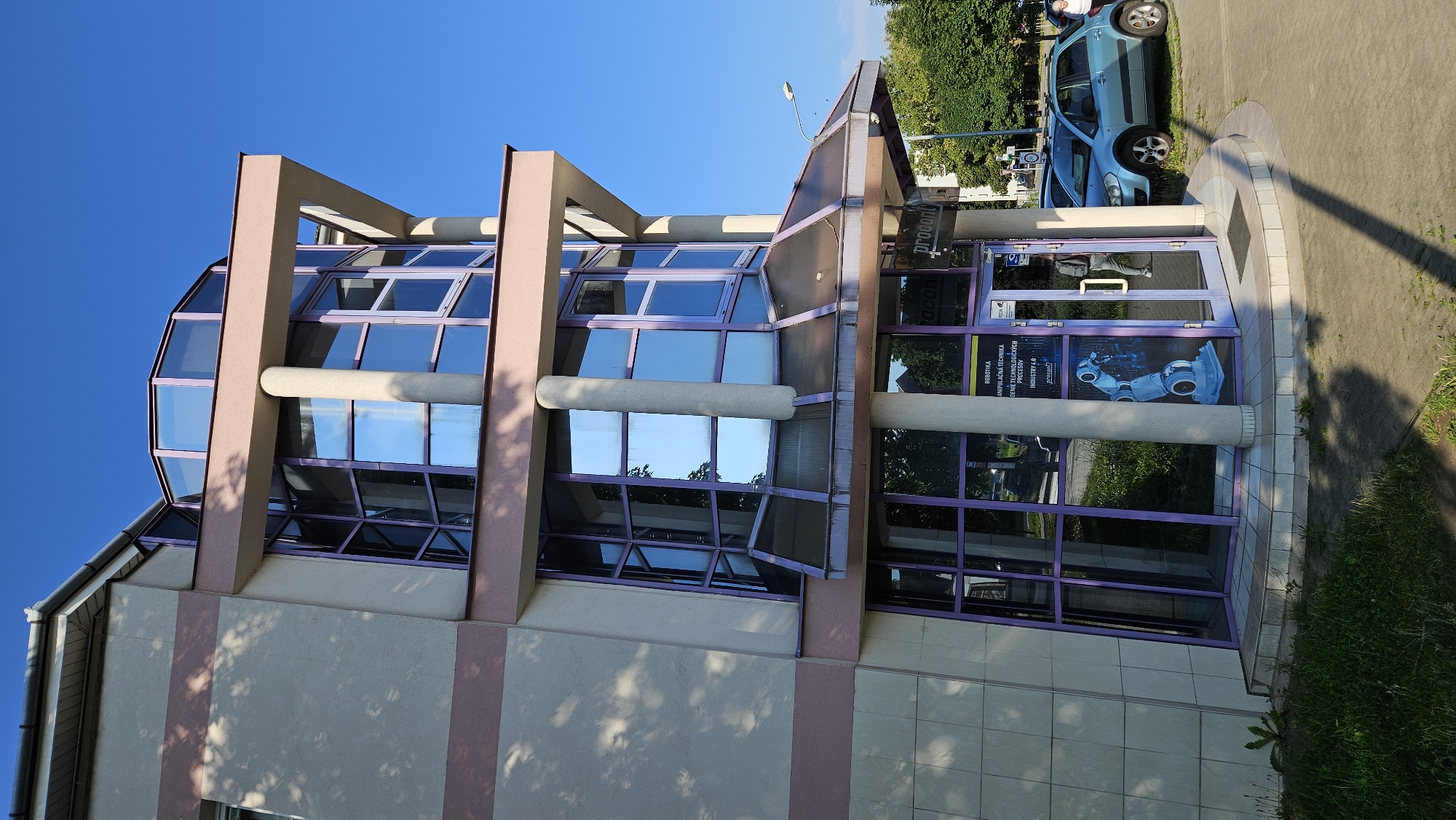 The image captures the exterior of a modern building with a curved glass facade, likely an office or commercial establishment, located in the city of Prešov, Slovakia. The building features a cream-colored exterior with horizontal pink bands. Its most striking feature is the multi-story, octagonal glass structure with purple-tinted frames, reflecting the blue sky and surrounding trees. A covered entrance area with a slanted glass roof is situated at ground level.
Through the glass entrance, a sign is visible that reads "ROBOTIKA MANIPULAČNÁ TECHNIKA RIADENÉ TECHNOLOGICKÝCH PROCESOV INDUSTRY 4.0" and "procont." A graphic of a blue industrial robot arm is displayed on the glass door.
In front of the building, a light blue car is parked on the paved area. A person, appearing to be a woman with light-colored hair, is walking away from the car towards the building. Another individual, seen through the glass entrance, is walking away from the building, carrying a backpack.
The immediate foreground consists of a paved walkway and a patch of green grass with some weeds. The background shows trees and other buildings, indicative of an urban setting. The scene is illuminated by natural sunlight, casting shadows on the ground. The sky is clear and bright blue.