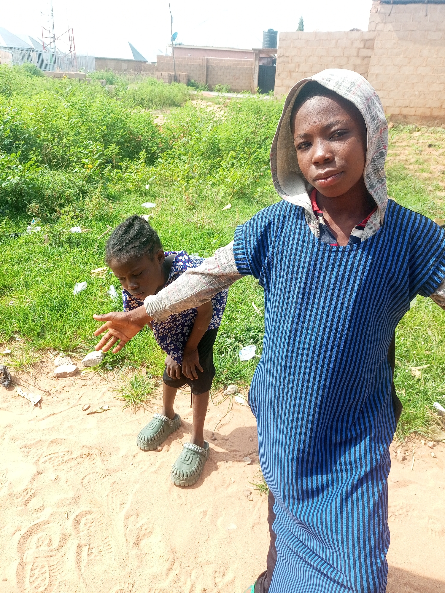 In Tempure, Nigeria, two young individuals stand on a dusty, sandy ground amidst dry vegetation and scattered debris. The person in the foreground, wearing a hooded blue and black striped shirt, looks directly at the camera with a neutral expression. Their hood is up, covering their head. To the left and slightly behind, another individual, presumably a child, is bent over, looking down at the ground with their left hand extended. They are wearing a blue and white patterned shirt and black shorts. The background reveals simple, light-colored brick buildings and what appear to be communication towers against a bright sky. The environment suggests an outdoor, possibly semi-urban or rural setting, with evidence of construction or development in the distance.