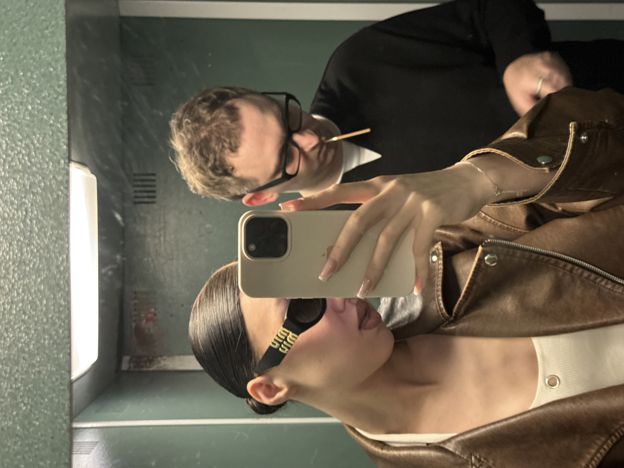 Two individuals are captured in a selfie taken in what appears to be an elevator. A woman with her hair slicked back, wearing a brown leather jacket over a white top and black sunglasses, holds a smartphone to take the picture. A man with short, curly hair and glasses is in the background, holding a thin stick-like object in his mouth. The elevator walls are green and textured, with a fluorescent light fixture visible at the top. The overall scene suggests a casual, everyday moment. The city of Khimki, Russia, is not depicted in the image.