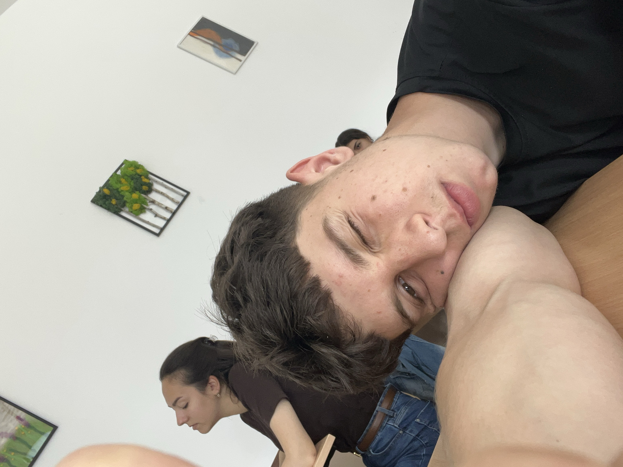 The image captures an indoor scene, likely in Zlatograd, Bulgaria, featuring a young man prominently in the foreground. He is oriented diagonally, resting his head on his bent left arm, which appears to be on a light wooden surface. He has dark, wavy hair, brown eyes, and freckles on his face. His expression is relaxed, with one eye partially closed, suggesting a casual or playful mood. He wears a black t-shirt and blue denim jeans with a brown belt.
In the background, against a plain white wall, other individuals and decorative items are visible. To the left of the main subject, a young woman with dark hair tied back is seen leaning forward, engaged in an unseen activity. Further back and to the right, part of another person's face is visible.
The white walls are adorned with framed artwork. In the upper right, a rectangular frame holds an abstract piece with shades of orange, blue, and brown. Below it, a square frame displays a three-dimensional decorative item resembling small green and yellow artificial trees or moss. A third framed piece, partially visible in the lower left, features green and possibly purple colors.
The lighting is bright and even, indicative of a well-lit indoor space. The scene appears to be a casual, candid moment among friends or acquaintances. No specific text is visible within the image.