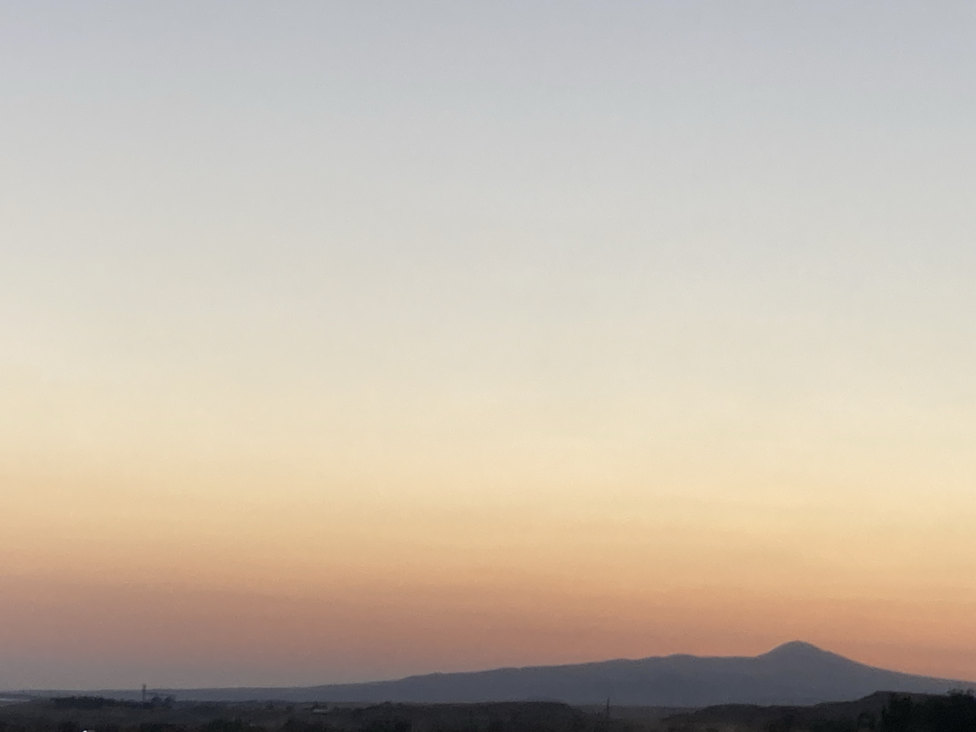 The image depicts a panoramic view of the sky over Avanos İlçe Merkezi, Turkey. The sky exhibits a gradient of colors, transitioning from a pale blue at the top to a warm orange and pink hue closer to the horizon, suggesting either sunrise or sunset. Silhouetted against this colorful sky is a mountain range, with a prominent conical peak in the center. The foreground shows a dark, indistinct landscape, possibly including some structures or natural formations, typical of the terrain around Avanos, which is known for its unique geological formations and proximity to mountains. No people or animals are visible in the frame. The overall impression is one of natural beauty and tranquility.