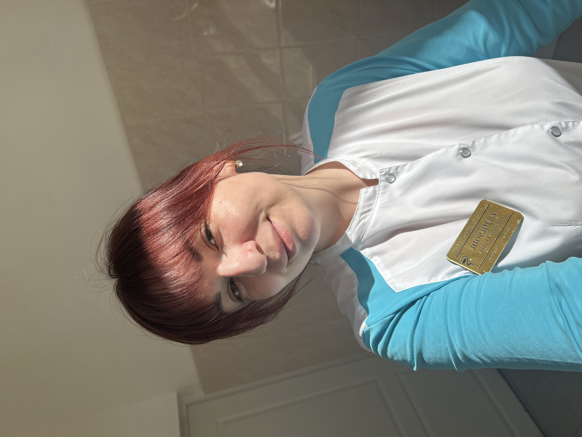 A woman with reddish-brown hair and bangs is shown in a medium shot, slightly angled, wearing a white lab coat with bright blue sleeves. She is wearing an identification badge that reads "ДЮРДІЄВА Дарья Сергіївна" and includes a caduceus symbol. The background is a light beige wall with a dark shadow cast on it, and a white door is partially visible at the bottom left. The photograph appears to have been taken indoors in Фоки, Russia.