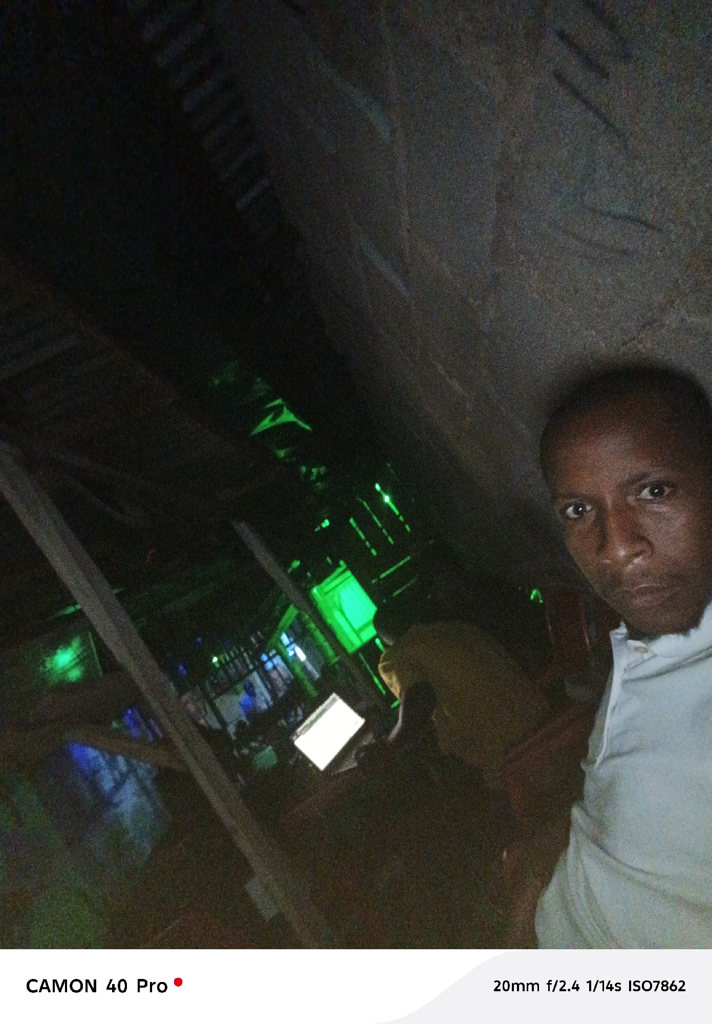 The image depicts a dimly lit interior space, likely a bar or lounge, with green ambient lighting. A man is visible in the foreground, positioned towards the right side of the frame, possibly taking a selfie. He is wearing a white shirt.
In the background, there are indistinct figures and objects. A laptop screen can be seen illuminated. The overall atmosphere suggests a nighttime setting in a commercial or entertainment venue.
The image includes the camera information "CAMON 40 Pro" and "20mm f/2.4 1/14s ISO7862".
There is no information that indicates the city of Jos, Nigeria.