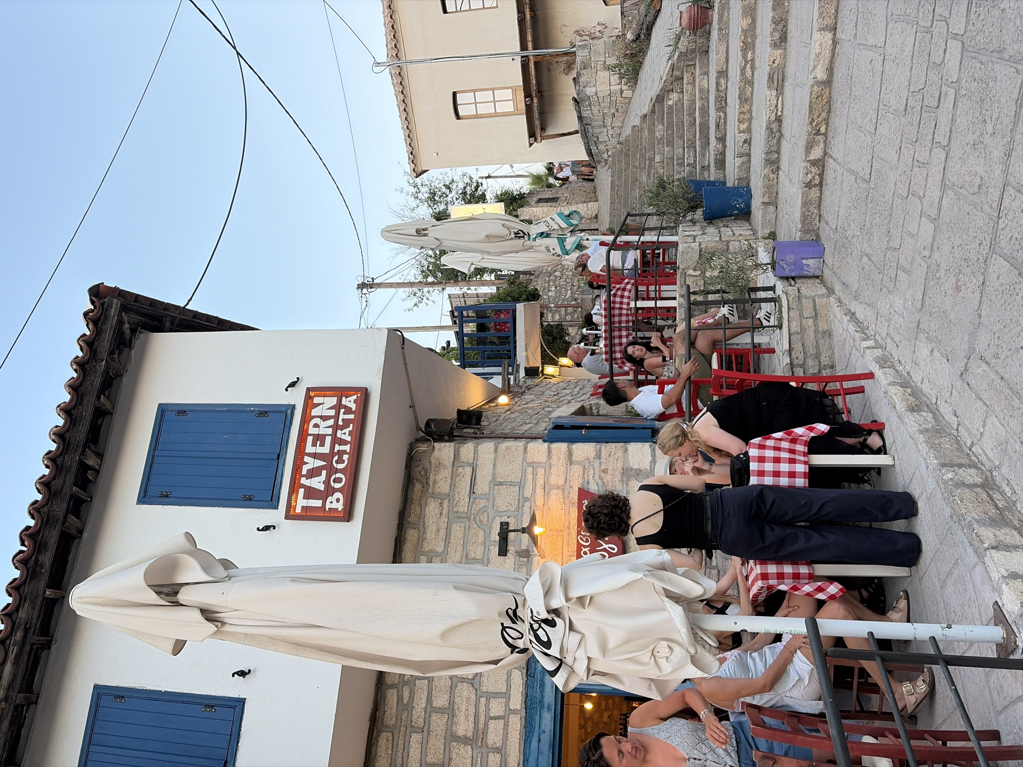 The image depicts an outdoor seating area of a tavern named "TAVERN BOGIATA" in Kassandra Municipal Unit, Greece. The tavern building features white walls, blue shutters on windows, and a red-lit sign. Several patrons are seated at tables covered with red and white checkered tablecloths. Some tables have umbrellas closed and tied, while others are open. The ground is paved with cobblestones and stone steps lead up to the right of the image, suggesting a hilly or terraced area. Various potted plants are visible, adding to the ambiance. The sky is clear and light blue, indicating it is daytime. People are engaged in dining and conversation, creating a relaxed atmosphere.
