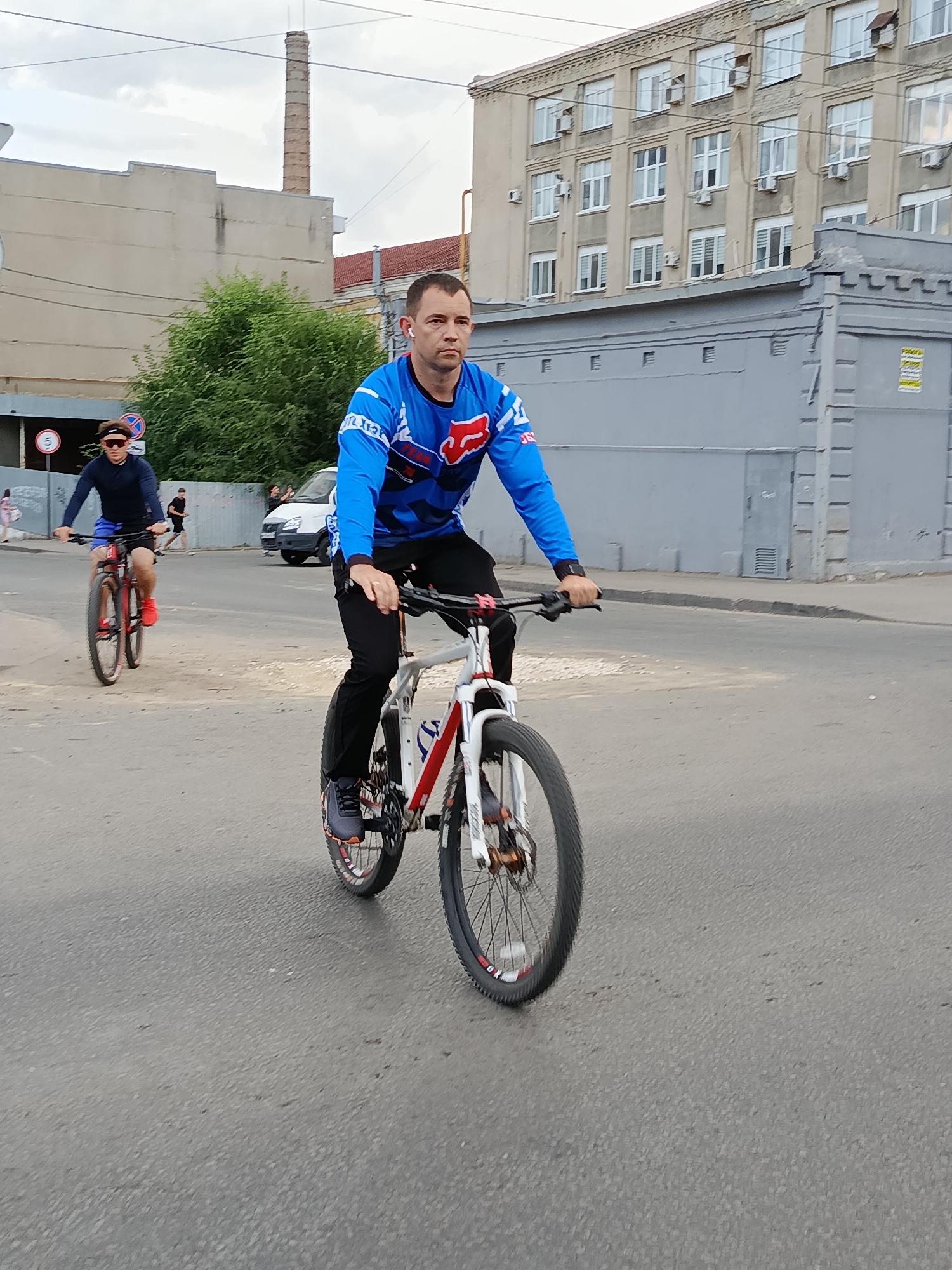 A man in a blue and white cycling jersey with red accents and black pants rides a white, red, and black mountain bike down a paved road in Saratov, Russia. He is wearing earbuds and looking forward. Another cyclist in a dark blue long-sleeved shirt and blue shorts, wearing a black headband and sunglasses, rides a red mountain bike in the background. The road is bordered by a sidewalk and buildings, including a light-colored building with many windows and air conditioning units, and a gray building with a decorative upper edge. A tall, industrial chimney is visible in the distance. The sky is overcast.