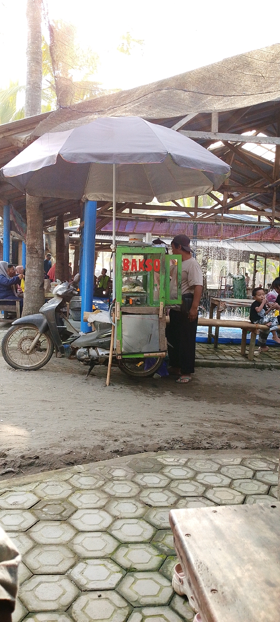 This image captures a street scene in Tambakmulyo, Indonesia, under a shaded area created by a large umbrella and a rustic overhead structure. The foreground features a tiled hexagonal pavement. The midground is dominated by a motorbike with a food cart attached to it. The food cart, labeled "BAKSO," is green and has glass panels displaying food items. A man in a dark t-shirt and black pants stands beside the cart, appearing to be attending to it.
To the left of the cart, several people are seated on benches under palm trees, including a woman wearing a hijab. In the background, more people are visible, some seated and others standing, suggesting a community gathering or marketplace. Further back, there is evidence of water, possibly a pond or pool, with a wooden railing. The overall environment appears to be an outdoor eating area or a casual public space with elements of local commerce and community life. The lighting suggests it is daytime, with sunlight filtering through the overhead structures and foliage.