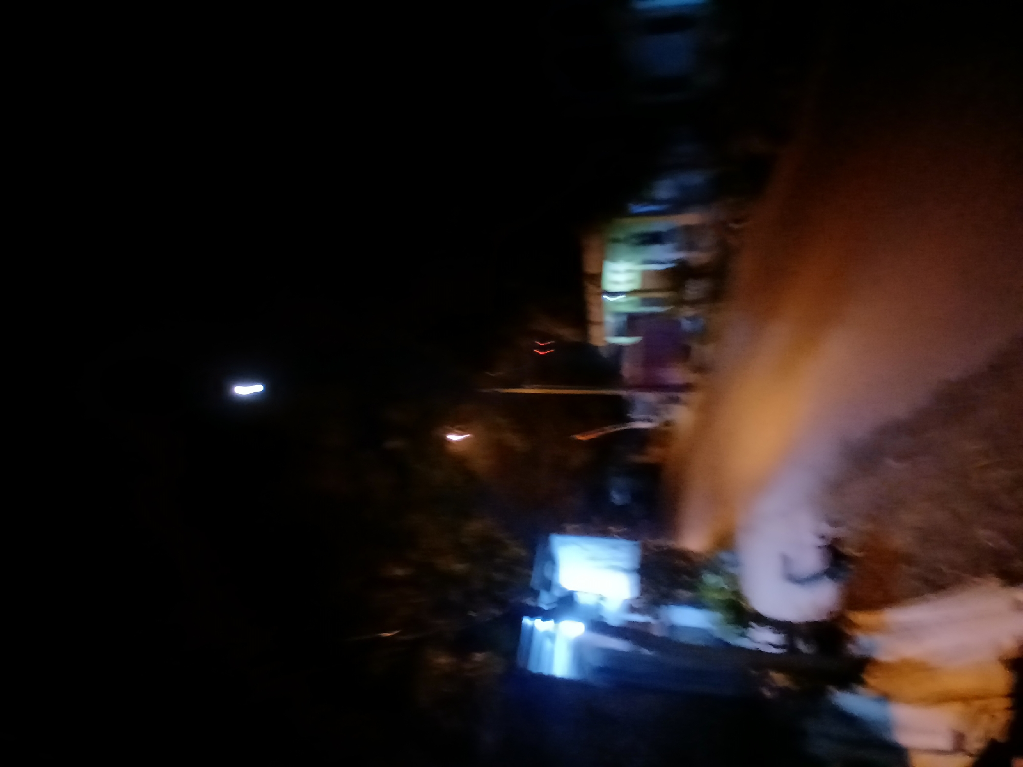 The image depicts a street scene at night in Kademangan Kulon, Indonesia. The scene is dimly lit, with artificial lights from buildings and street lamps creating streaks and blurs due to motion or low light conditions. On the left side of the image, a building with illuminated windows is visible, with blueish light emanating from within. A street with a reddish-orange hue runs through the center of the frame, suggesting it is paved or covered in dirt reflecting ambient light. Trees and foliage are present in the background, appearing dark and indistinct. Further down the street, other structures are faintly visible, some with illuminated windows or overhead lights. A single, bright, vertical streak of white light is prominent in the upper portion of the sky, possibly a light pole or a distant light source. The overall impression is of a quiet, residential street at night, with the limited visibility creating a somewhat abstract and impressionistic view.