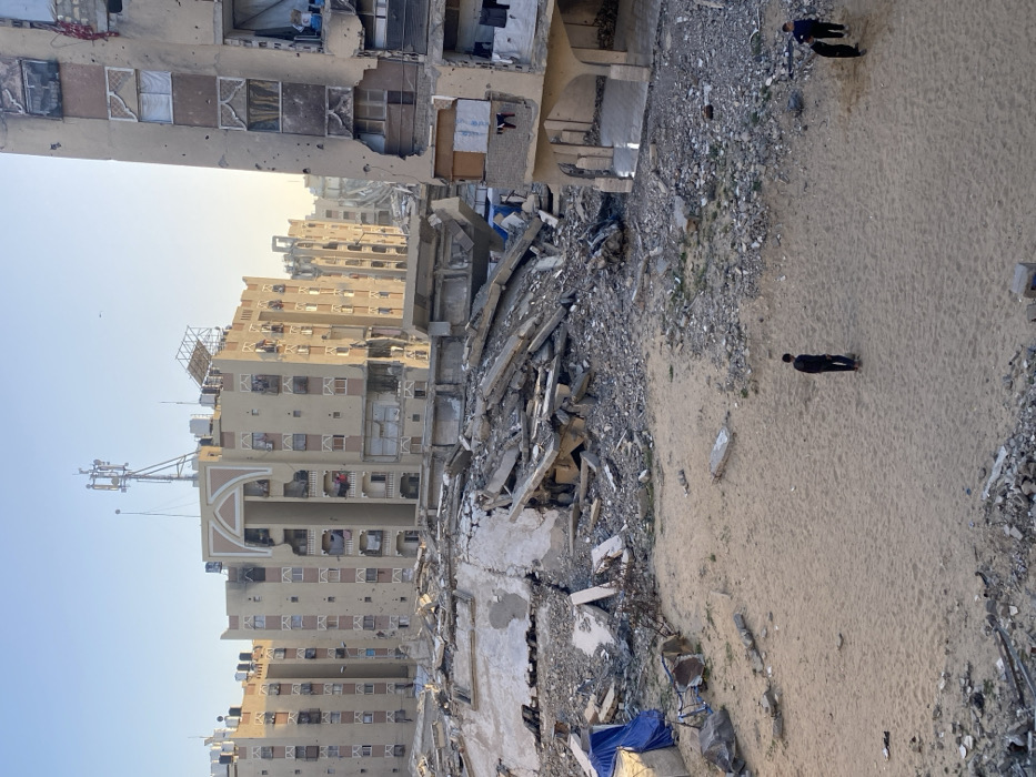 The image presents a desolate urban landscape marked by extensive destruction. Multiple multi-story buildings, constructed primarily of concrete, dominate the scene. Many of these structures exhibit severe damage: windows are shattered or missing, exposing dark interiors; sections of walls have collapsed, revealing rebar and debris; and facades are peppered with small, dark pockmarks, consistent with bullet holes or shrapnel impacts.
In the center of the image, a massive pile of rubble, composed of concrete slabs, twisted rebar, and pulverized building materials, signifies the complete collapse of one or more structures. This debris stretches across the mid-ground, separating the more intact buildings from a large, sandy expanse in the foreground and to the right. The ground in this sandy area is light brown and appears mostly barren, though scattered pieces of smaller debris and gravel are visible. A blue tarp or fabric lies near the edge of the rubble pile closest to the sandy ground.
Three individuals, all appearing to be adult males dressed in dark clothing, are present in the sandy area. One man stands alone in the middle of this open space, facing away from the camera, seemingly observing the devastation. Further to the right, closer to the rubble, two other men are seen; one appears to be standing, while the other is less distinct, possibly bent over or sitting. Their presence suggests they are surveying the aftermath of the destruction.
The sky above is clear and light blue, indicating fair weather. The sunlight casts soft shadows and has a slightly warm hue, suggesting either early morning or late afternoon. On the roof of one of the prominent, taller buildings in the mid-ground, a large antenna or cell tower structure is clearly visible, along with multiple external air conditioning units on its windows.
The overall scene conveys a profound sense of devastation and the quiet aftermath of a destructive event, likely a conflict or disaster. There is no visible text in the image.