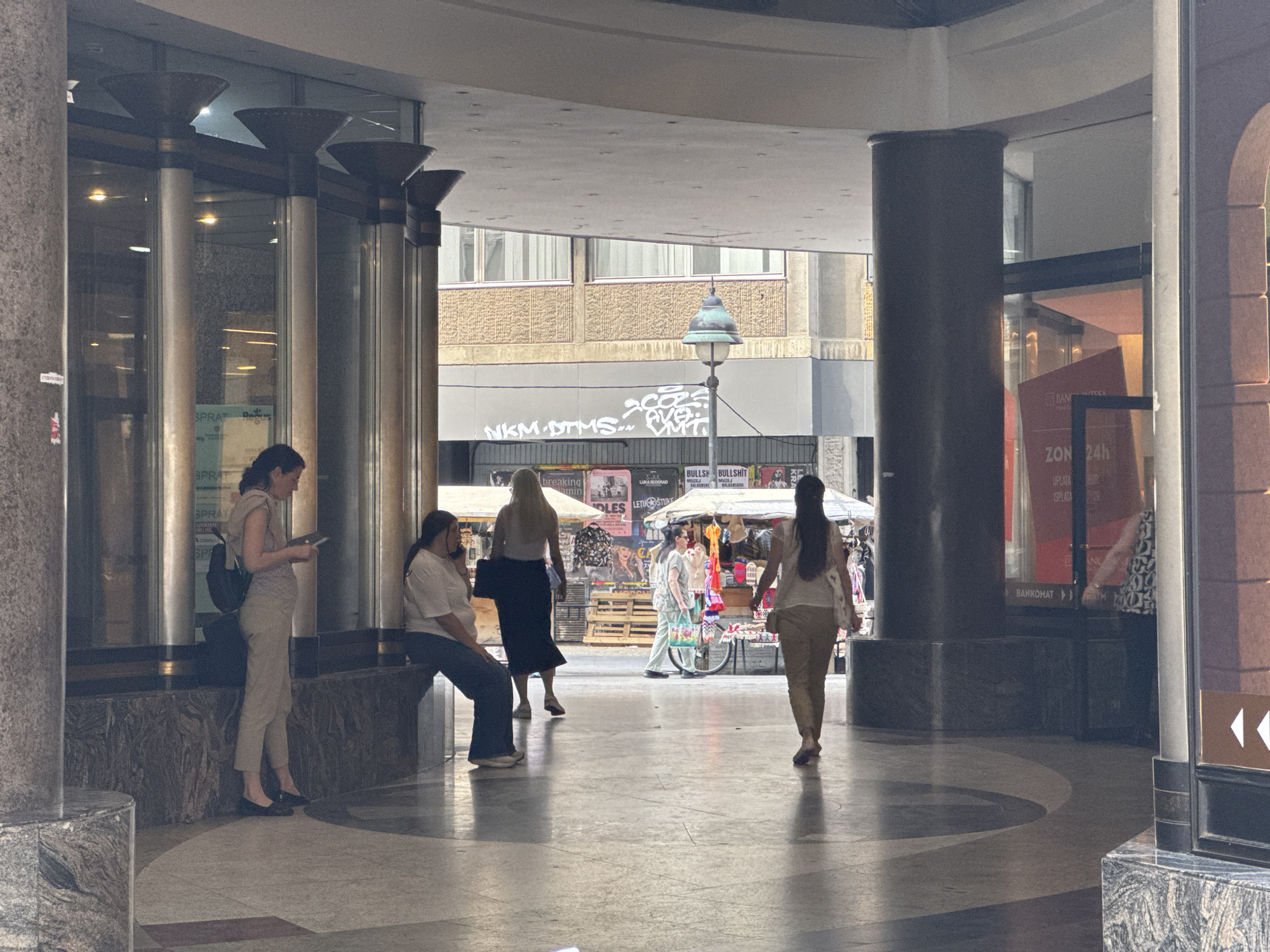 The image depicts an outdoor walkway or corridor in Belgrade, Serbia, characterized by architectural columns and shops. In the foreground, a woman stands looking at her phone, dressed in light-colored pants and a t-shirt, with a backpack. To her left, another person is seated on a bench, facing away from the camera. Further into the corridor, several individuals are visible, some walking and others browsing stalls that appear to be selling clothing and accessories. The stalls are set up under canopies. In the background, a building with a textured facade and windows is visible. Sunlight casts distinct shadows on the ground. The overall scene suggests a public space with commercial activity.