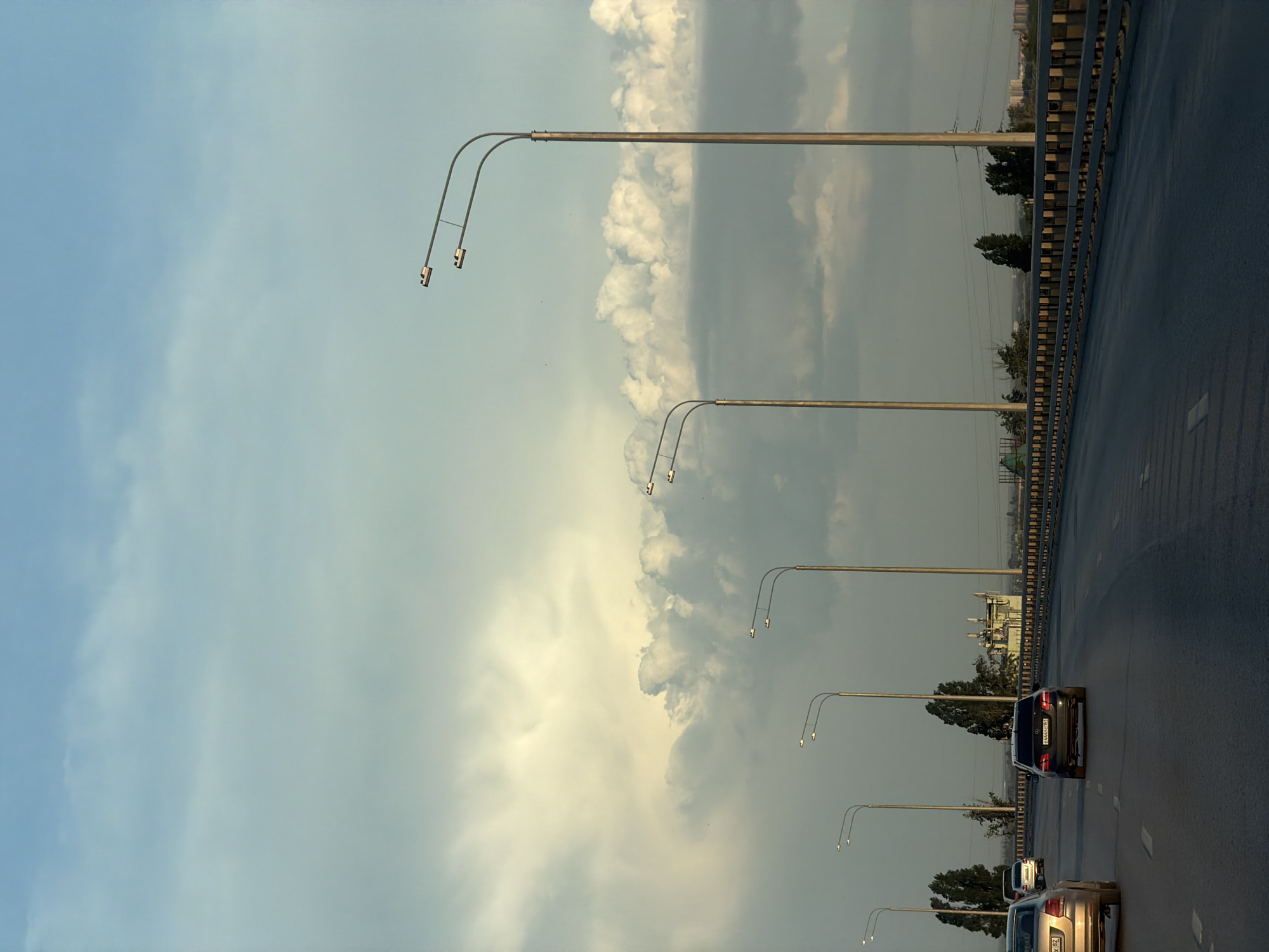 The image depicts a highway in Rostov-on-Don, Russia, with several cars traveling on the road. The sky above is filled with clouds. The highway is lined with streetlights on poles. A bridge's guardrail runs along the side of the road. Buildings and trees are visible in the background.