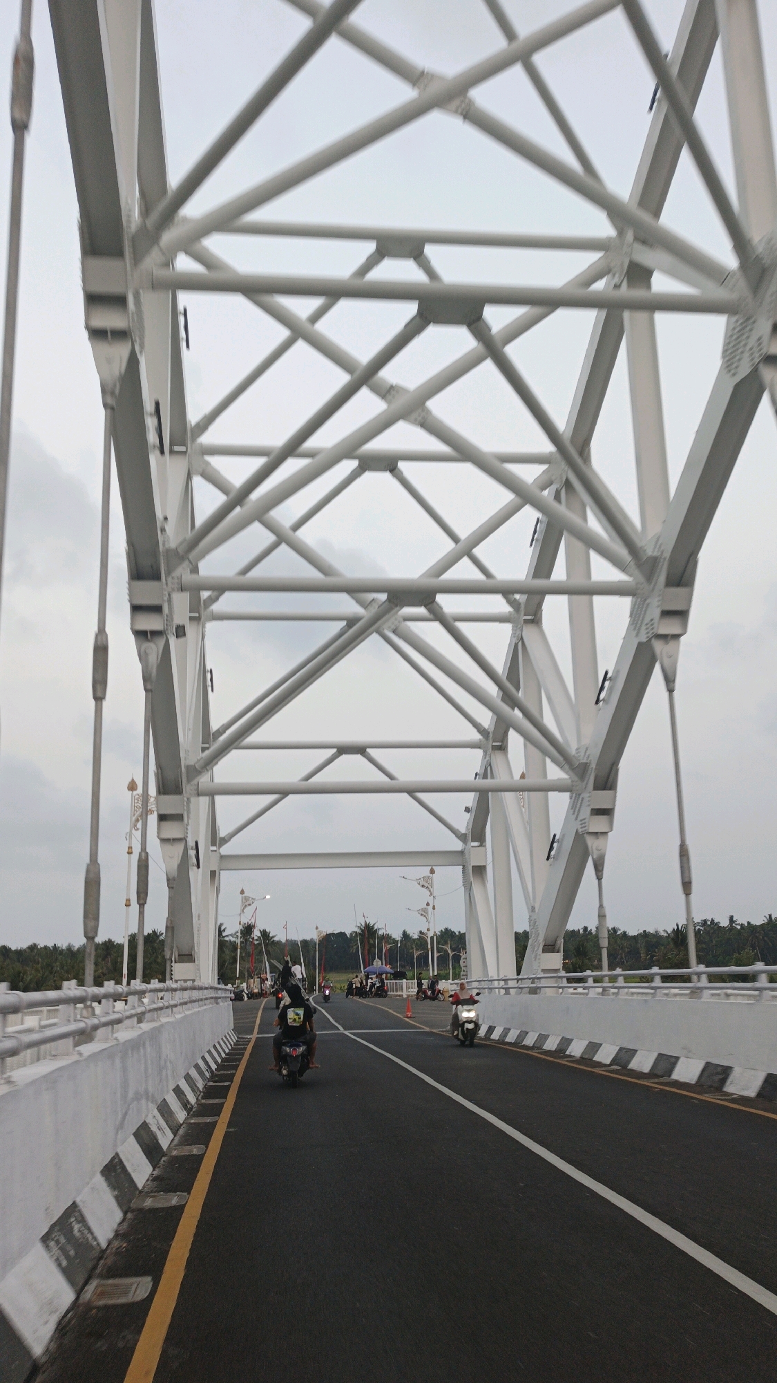 The image shows a view from a vehicle driving across a modern, white steel truss bridge in Desa Cijulang, Indonesia. The bridge has a wide asphalt road with clear white lane markings and a yellow line bordering the left side. A concrete barrier with black and white diagonal stripes runs along the left edge of the road.
Several motorcycles are present on the bridge. In the foreground, a motorcycle with two riders is moving away from the camera. To the right, another motorcycle is also traveling in the same direction. In the mid-ground, a cluster of motorcycles and people are stopped or gathered on the right side of the road, near decorative poles and flags. Further in the distance, more motorcycles and a few vehicles are visible, suggesting a moderate amount of traffic.
The sky is overcast with a light gray hue. On either side of the bridge, beyond the railings, there are glimpses of lush green trees and foliage, indicating a rural or semi-rural setting. The overall scene depicts a functional bridge in use by local traffic in Indonesia.