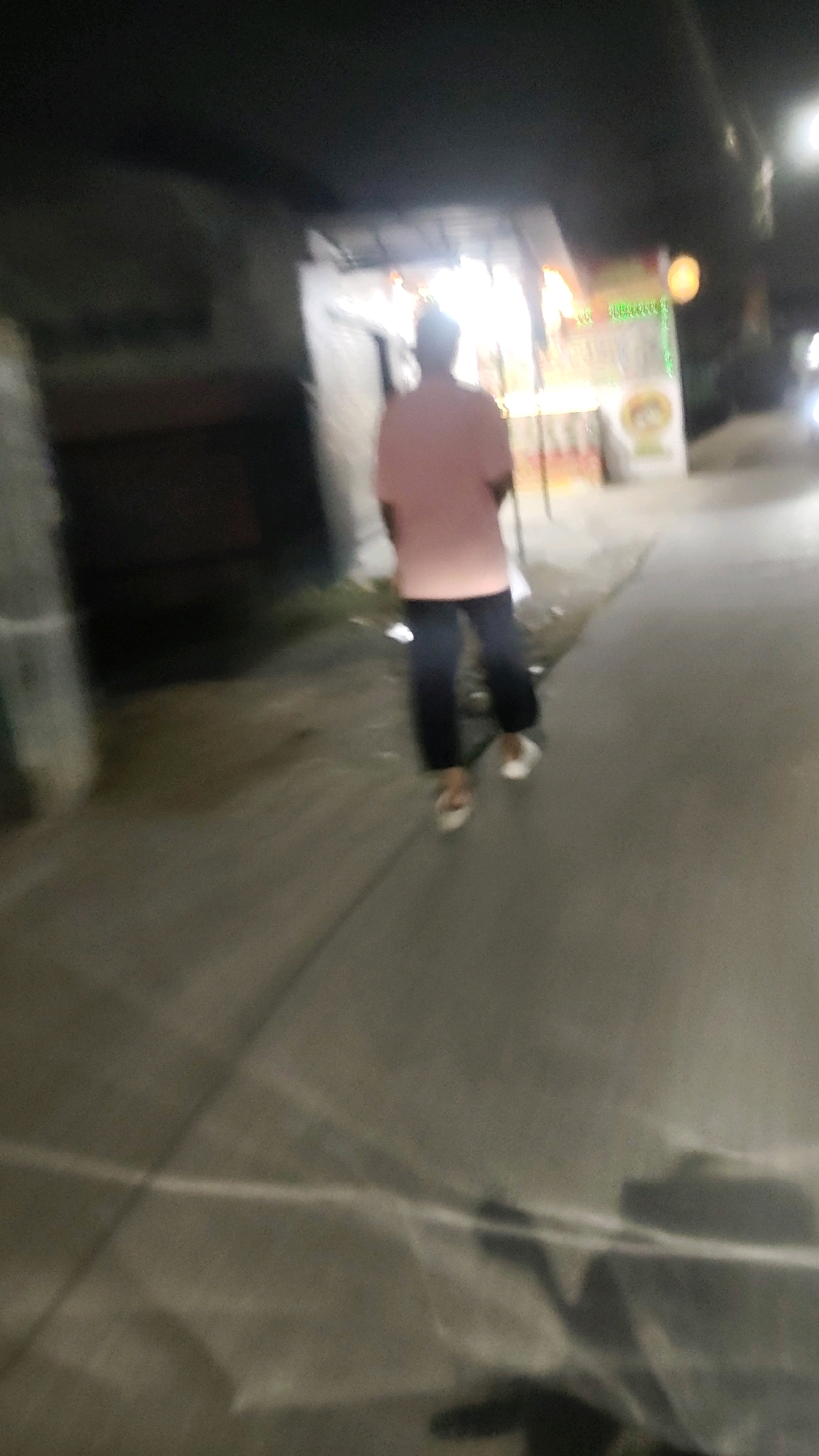 A person wearing a pink shirt and dark pants walks away from the camera on a sidewalk at night in Mekarmukti, Indonesia. The person is walking towards a brightly lit food stall that is open for business. The food stall has a colorful sign and is illuminated by string lights. The sidewalk is made of concrete and is wet from a recent rain. The street is lined with trees and buildings. The scene is dimly lit, with the primary light source coming from the food stall. The image is slightly blurry, suggesting it was taken with a handheld camera in low light conditions.